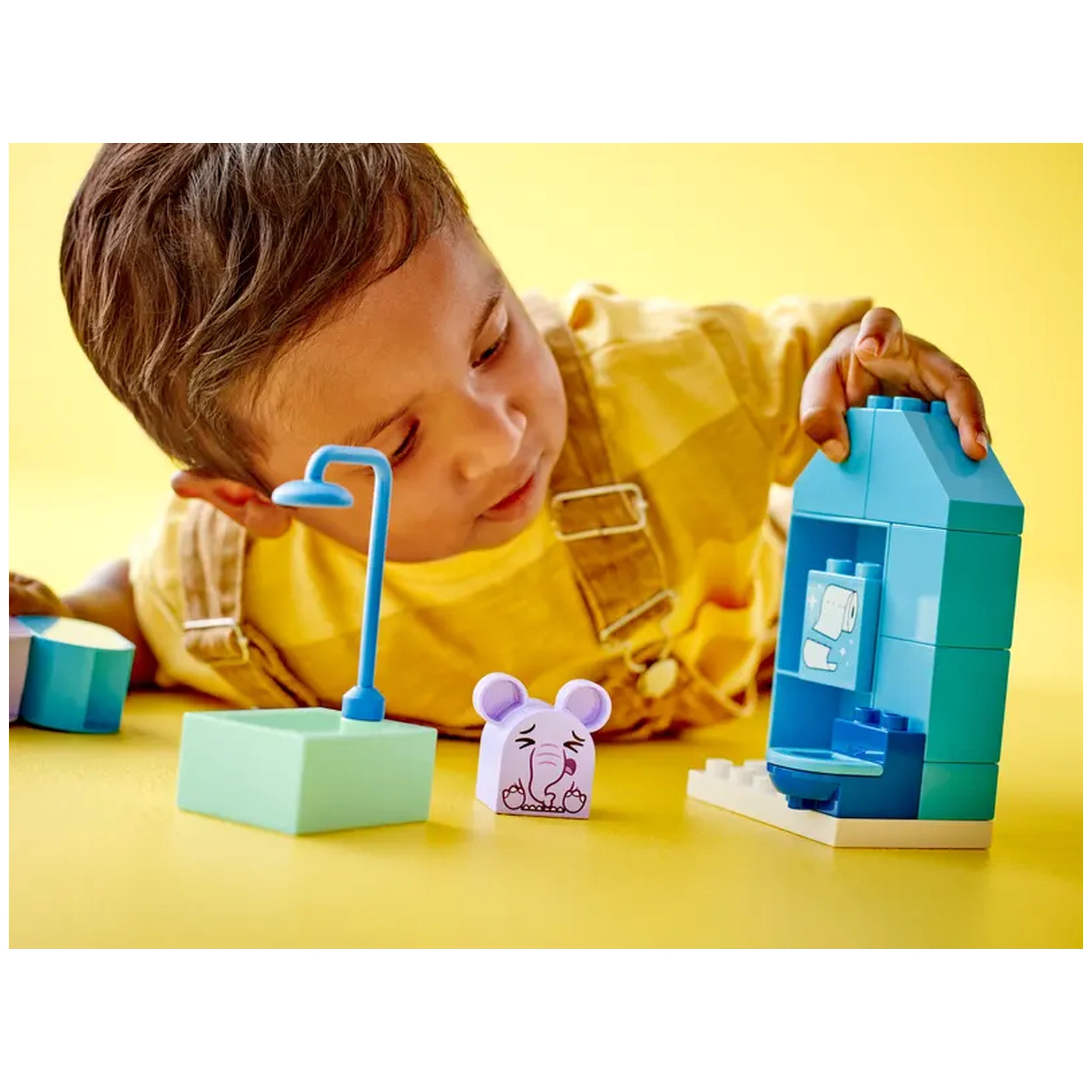 A young child in a yellow shirt plays with a colorful LEGO® DUPLO® bath set, featuring a teal bathroom setup, a blue shower, and a light green tub, alongside an adorable purple elephant figure, on a bright yellow surface.