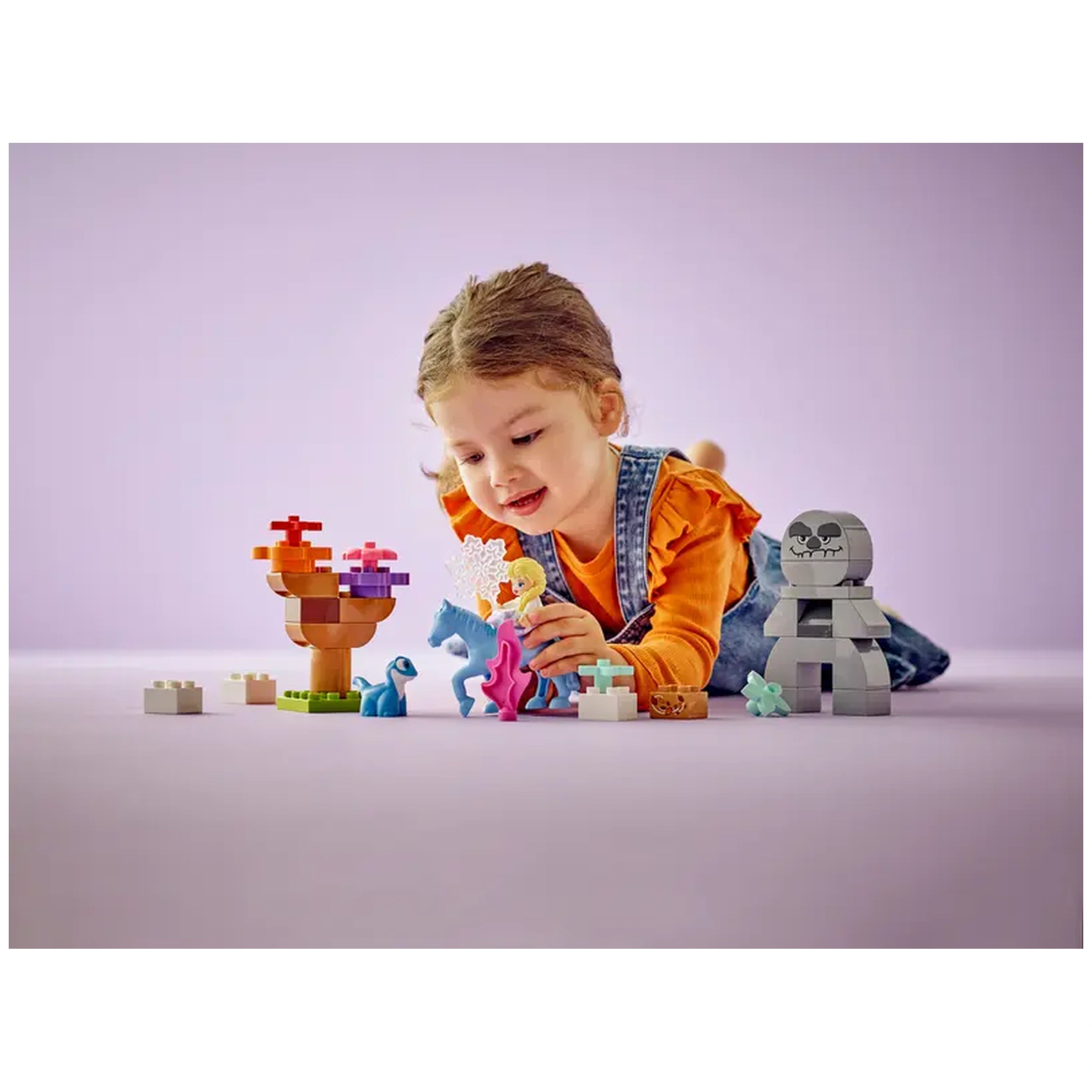 A young girl in a bright orange shirt plays on a soft purple surface, interacting with colorful LEGO® DUPLO® toys. She is surrounded by a buildable tree, a blue dinosaur, a pink horse, and figures of Elsa and Bruni, engrossed in imaginative play.
