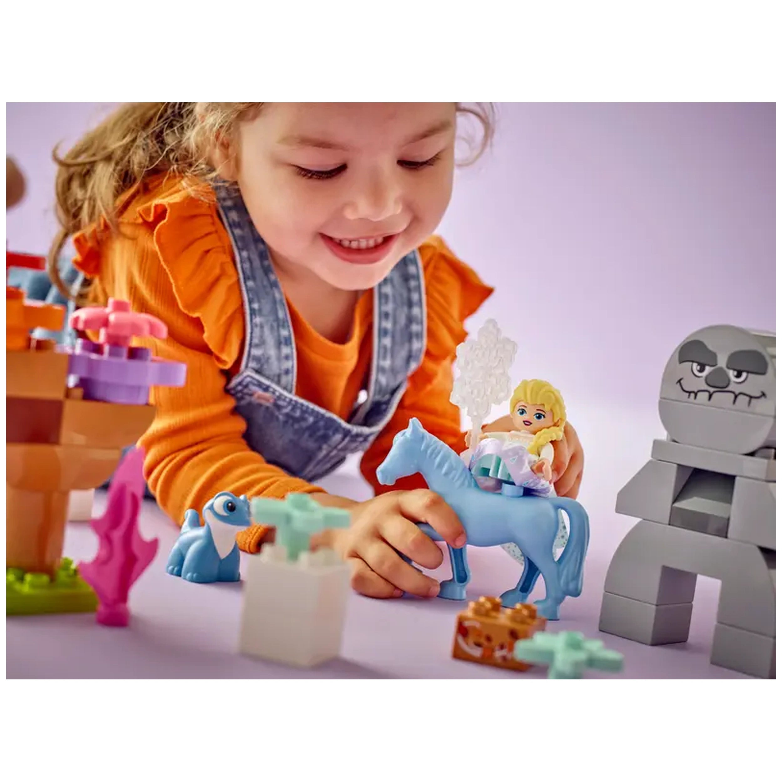 A young girl in an orange shirt smiles while playing with LEGO® DUPLO® figures, including Elsa on a blue horse, surrounded by colorful blocks and character toys.