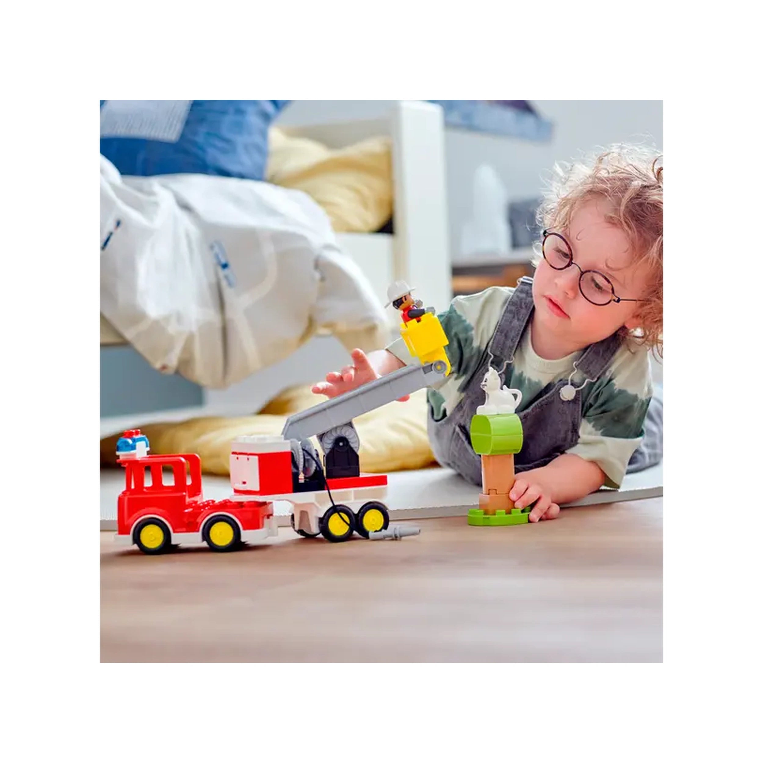 A young child with curly hair and glasses plays on the floor with a colorful LEGO® DUPLO® fire truck construction set, featuring a movable ladder and a tree with a toy cat. The child interacts with the truck while demonstrating imaginative play. A cozy bedroom is visible in the background, with soft bedding and pillows.