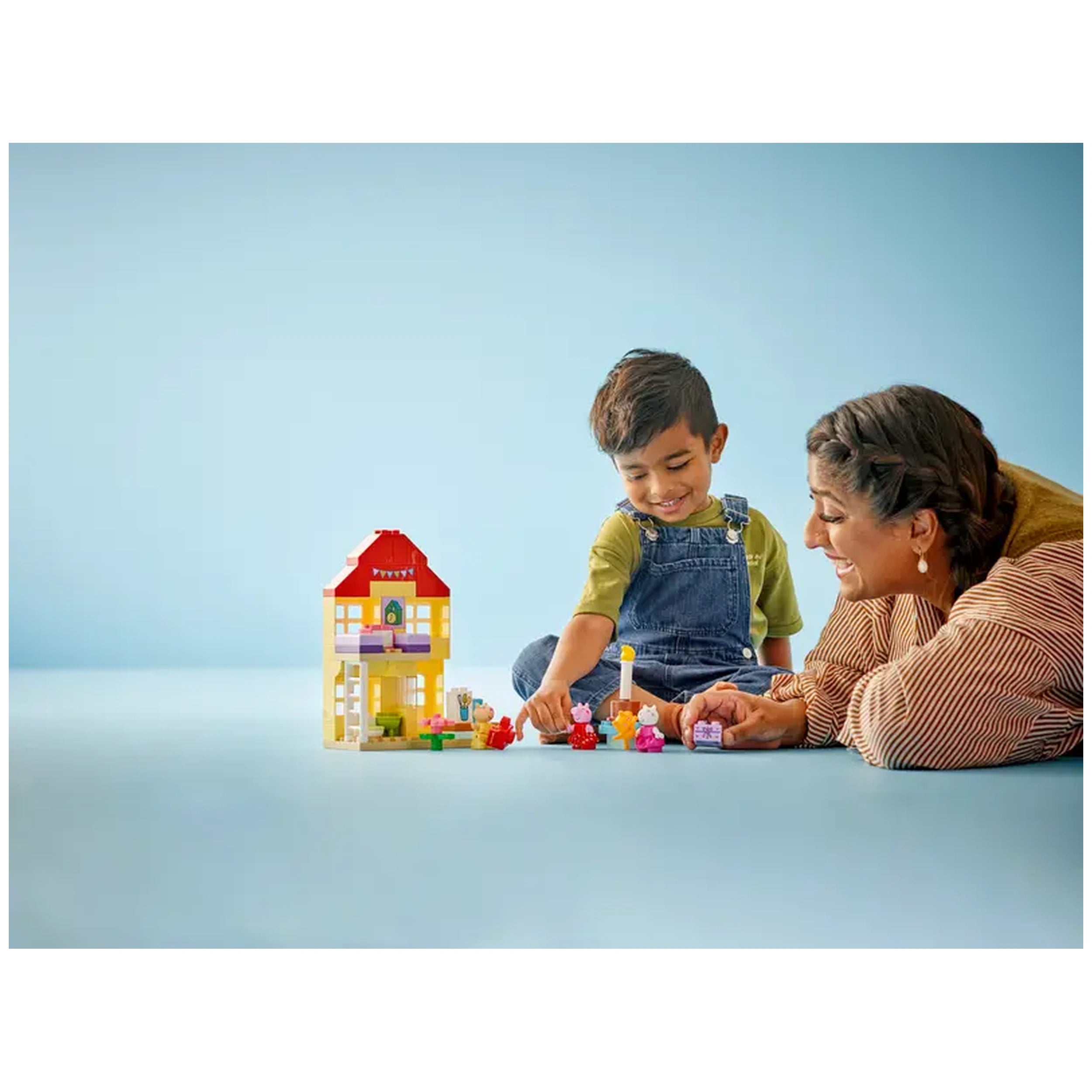 A young boy and a woman happily play with the LEGO® DUPLO® Peppa Pig Birthday House set, arranging colorful figures in front of the house.