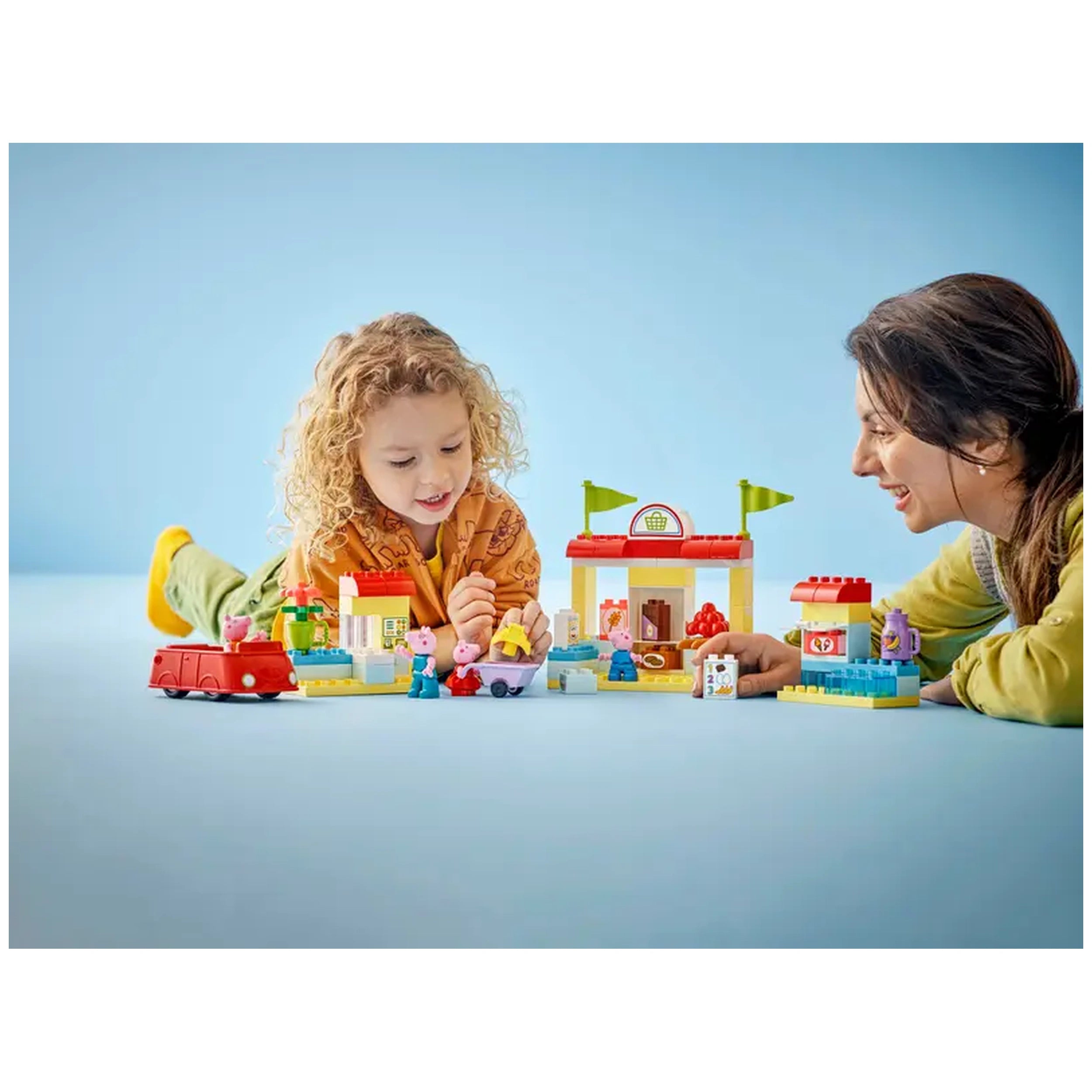 A child and an adult play together with the LEGO® DUPLO® Peppa Pig Supermarket set, featuring colorful toy buildings and familiar characters. The child laughs while interacting with Peppa Pig figures and a red toy car, set against a light blue background.