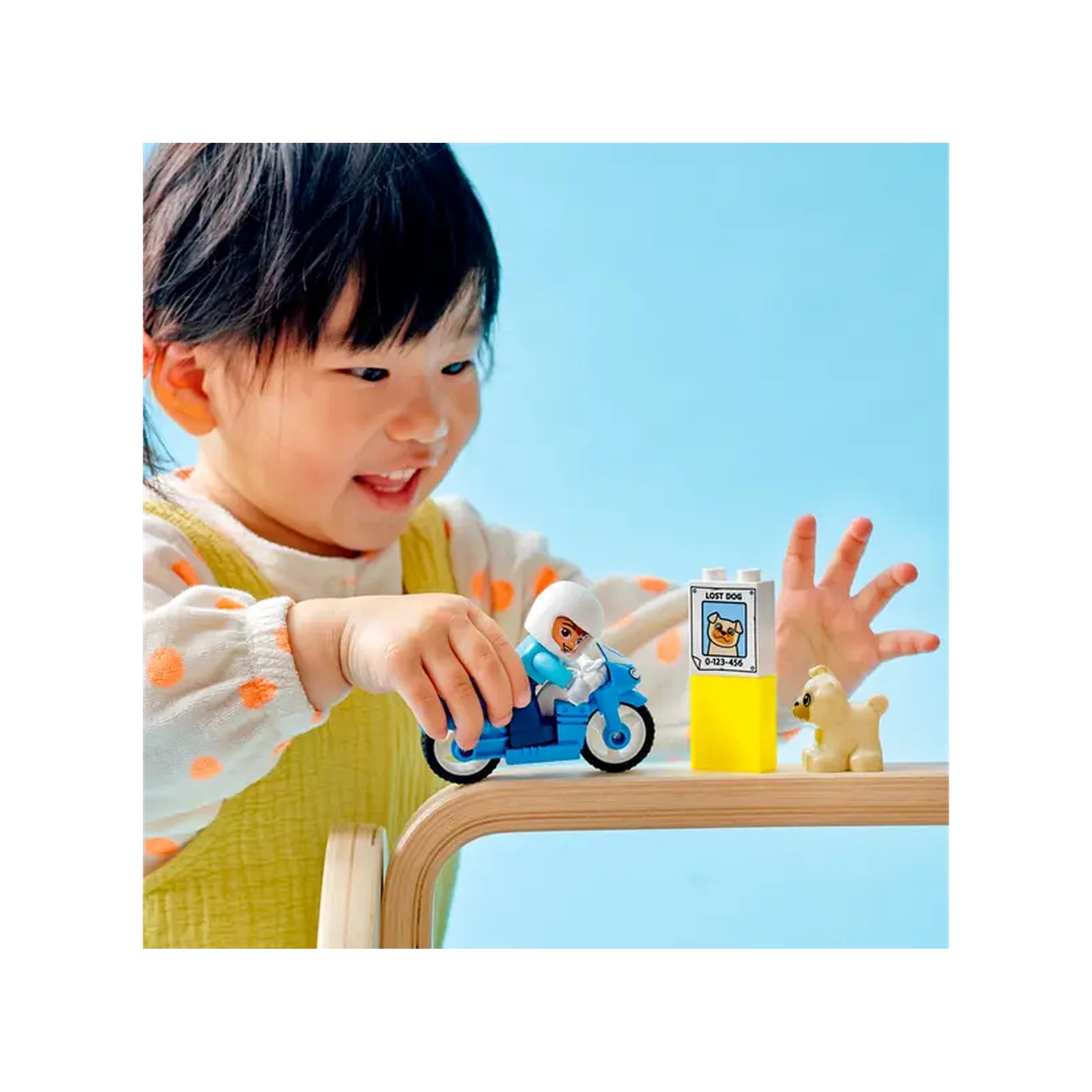 A young child joyfully plays with a blue LEGO® DUPLO® motorcycle, featuring a police officer figure, next to a playful dog and a 'lost dog' poster on a wooden table. The background is a soft blue, enhancing the vibrant colors of the toys and the child's outfit.