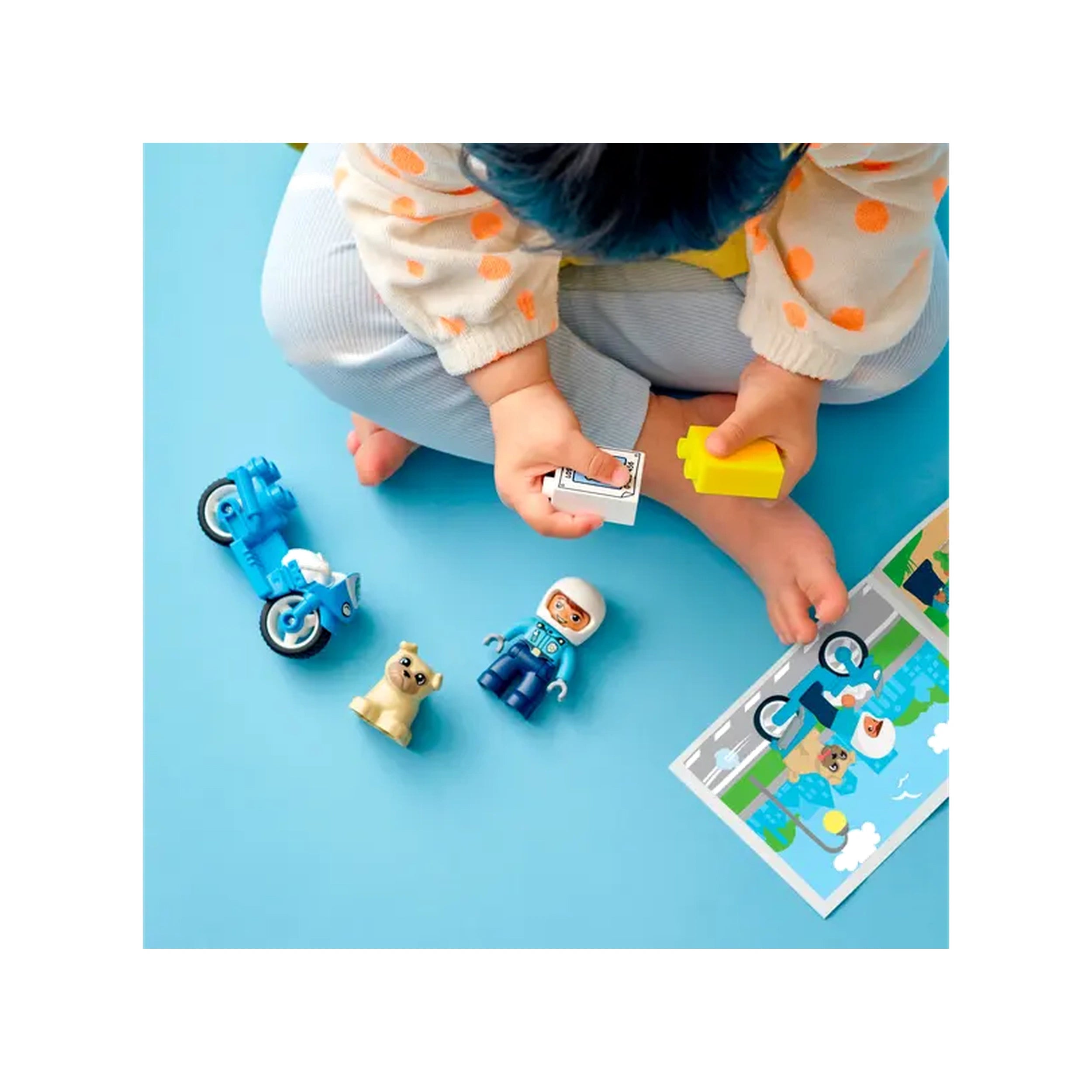 A child plays on a blue background with LEGO® DUPLO® Rescue Police Motorcycle set, holding a yellow block and a poster while surrounded by a police officer figure, a dog figure, and a blue motorcycle.