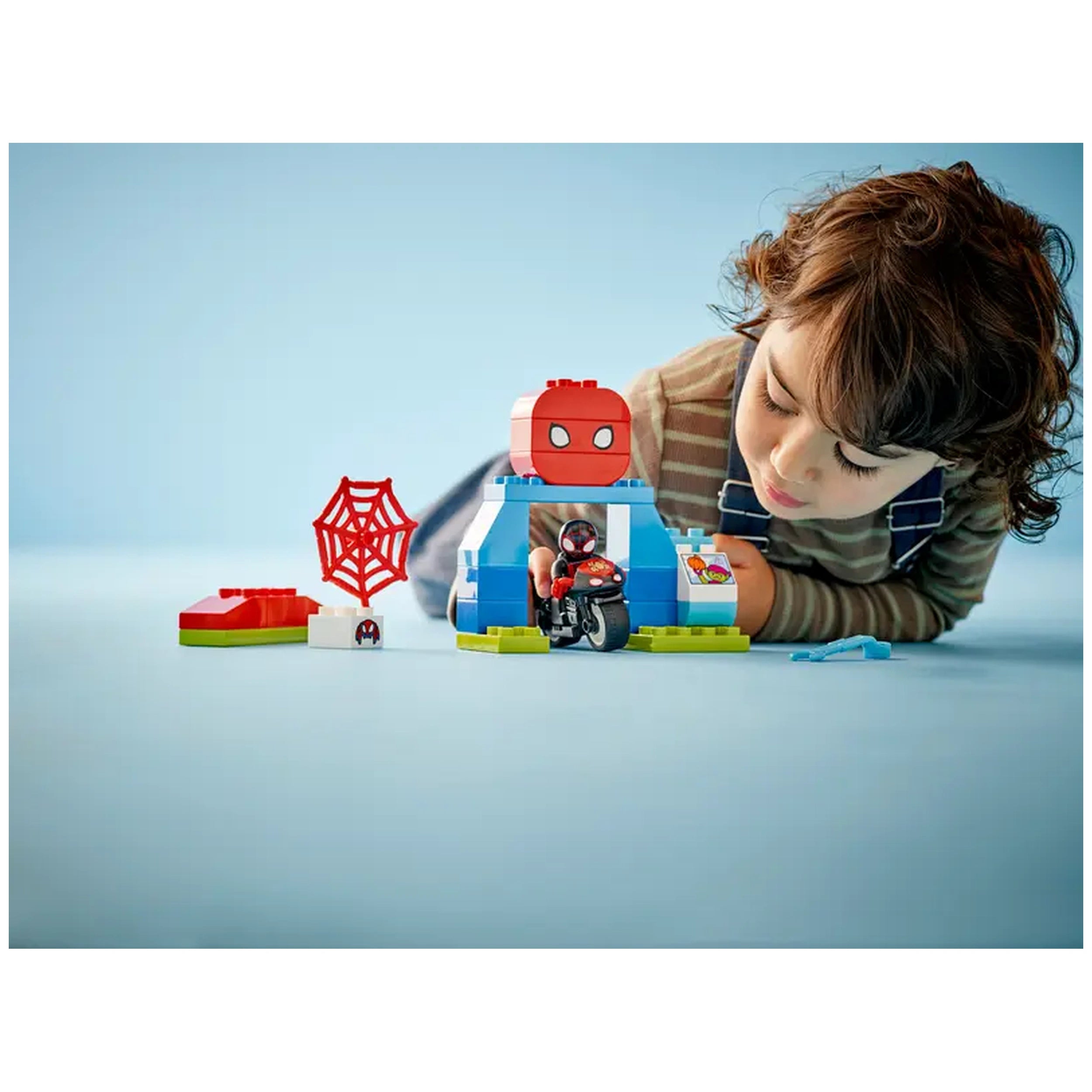 A child lies on their stomach, building with a LEGO® DUPLO® set featuring a Spider-Man figure, motorcycle, and colorful blocks.
