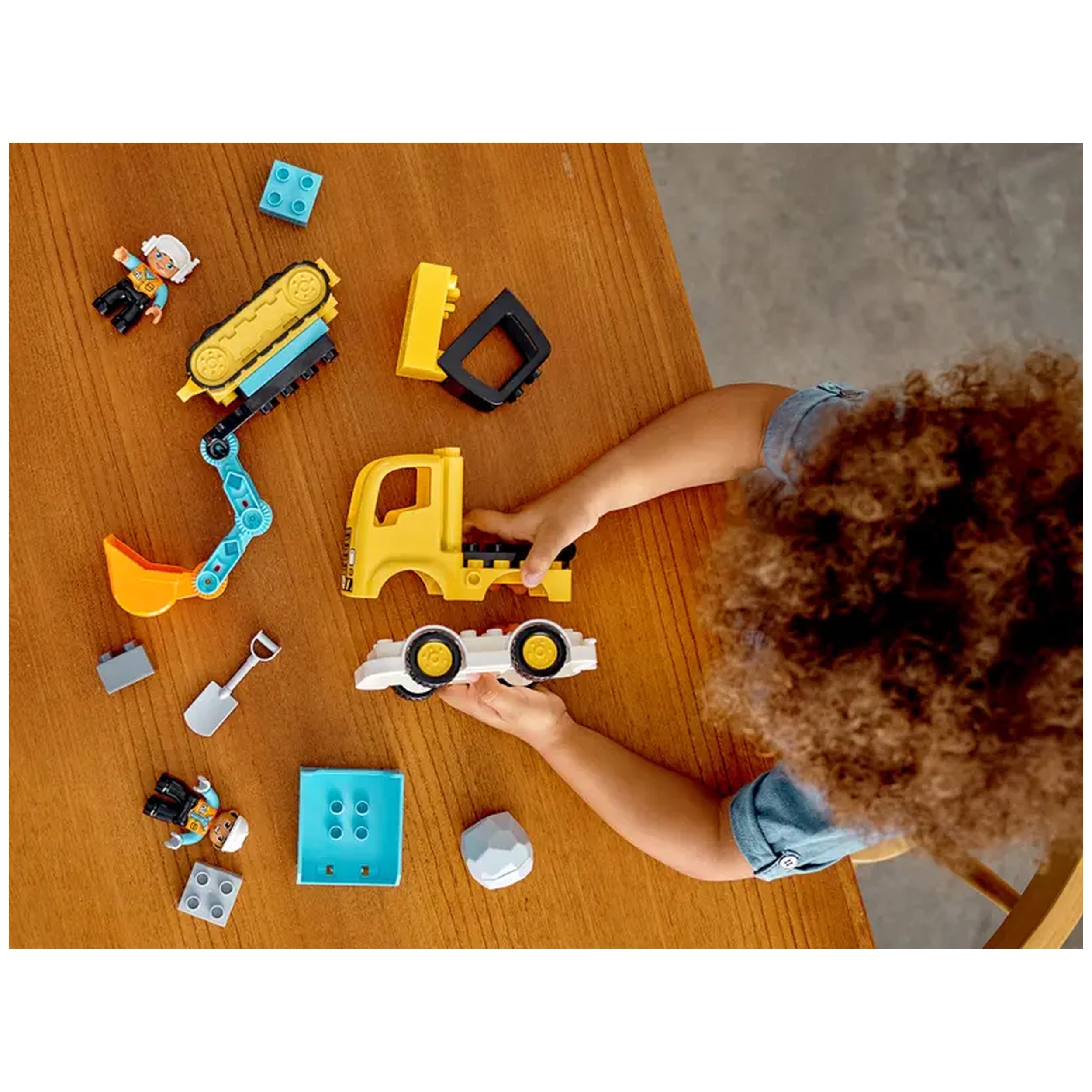 A toddler with curly hair plays with a yellow truck and a white vehicle on a wooden table, surrounded by colorful construction toys and a construction worker figure.