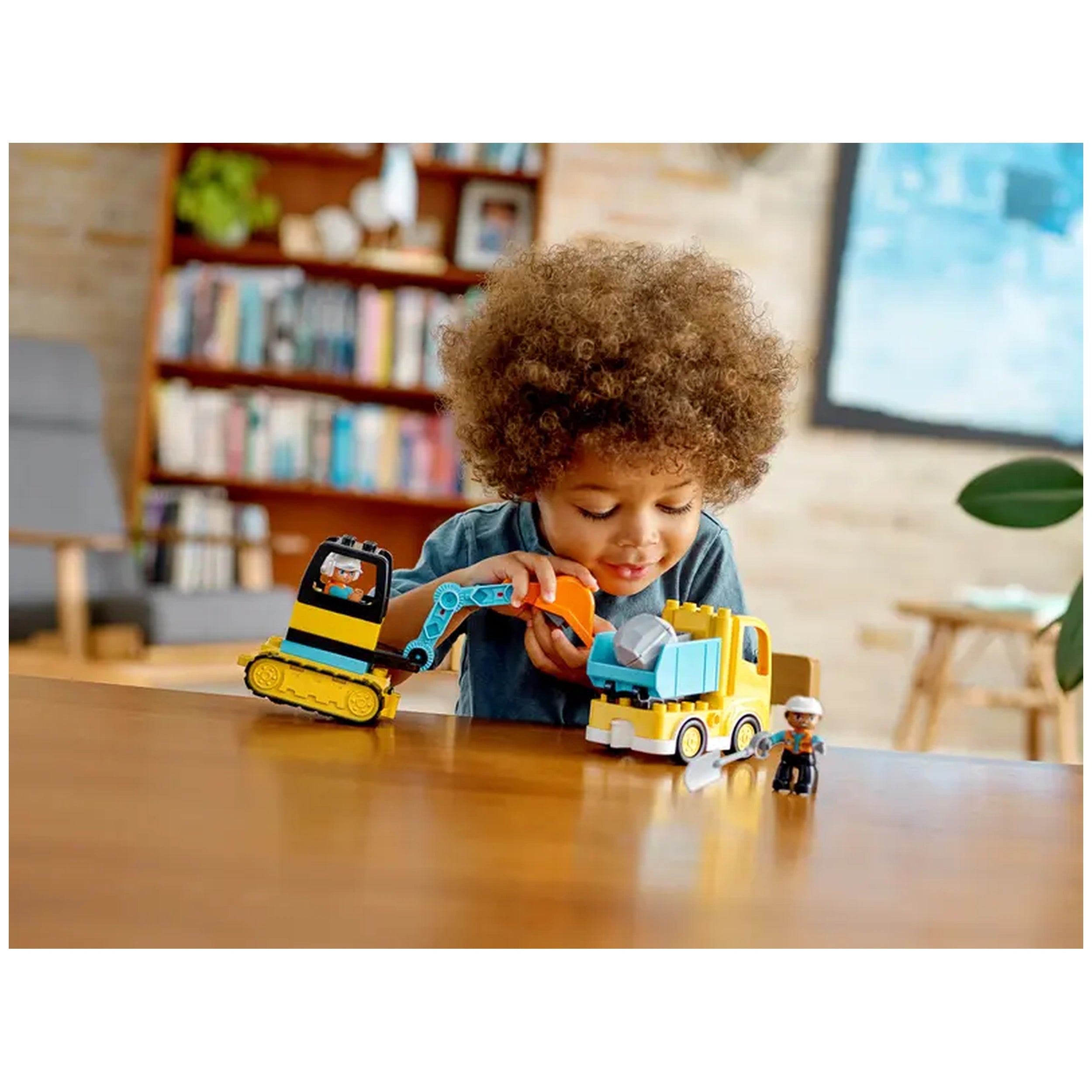 A child plays with a colorful LEGO® DUPLO® construction set, manipulating a blue truck and a yellow excavator on a wooden table, with a smiling construction-worker figure nearby.