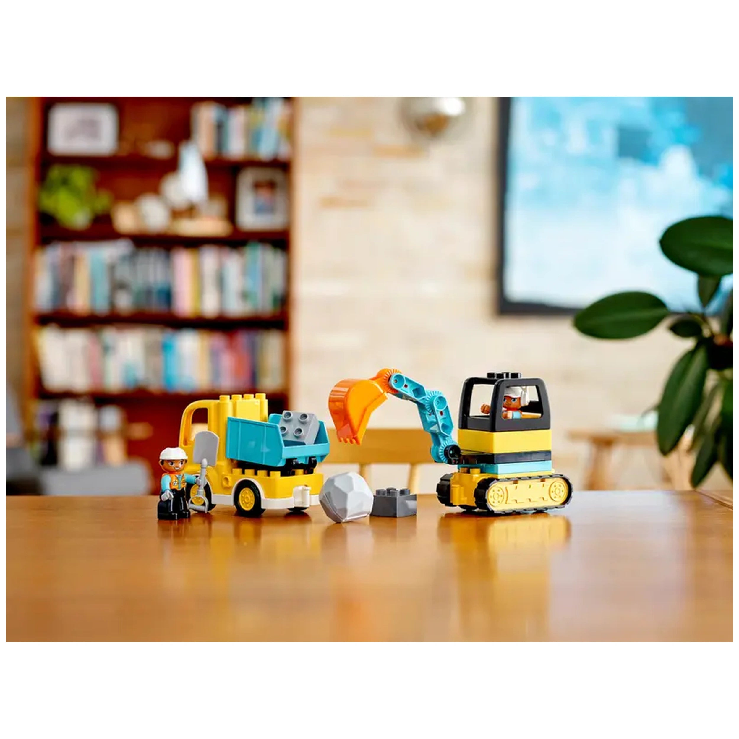 LEGO® DUPLO® construction set featuring a yellow truck and a tracked excavator, with two construction-worker figures and building blocks on a wooden table.