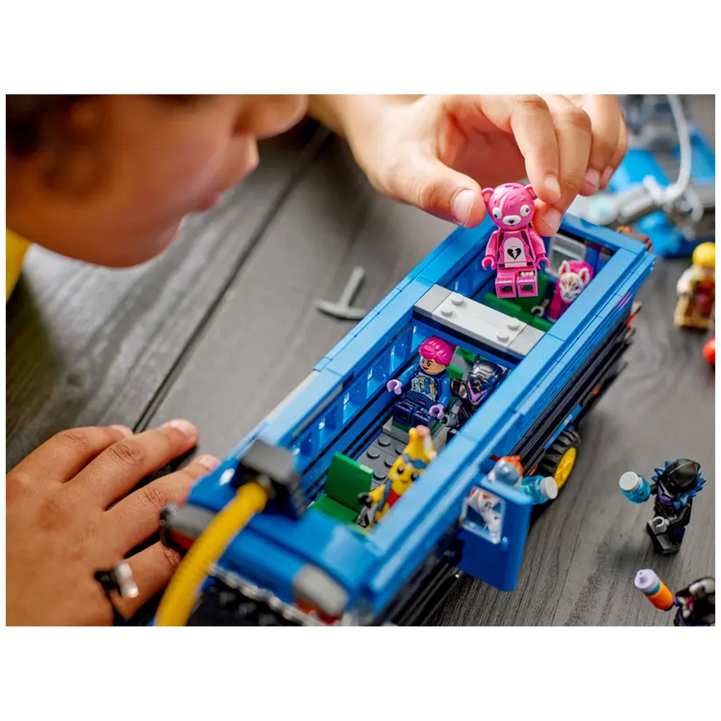 A child intently plays with the LEGO Fortnite Battle Bus, holding a pink character figure above. Inside the bus, colorful mini-figures are seated, showcasing vibrant accessories. The blue bus has a detailed design, with green seating and a removable roof, situated on a wooden surface.
