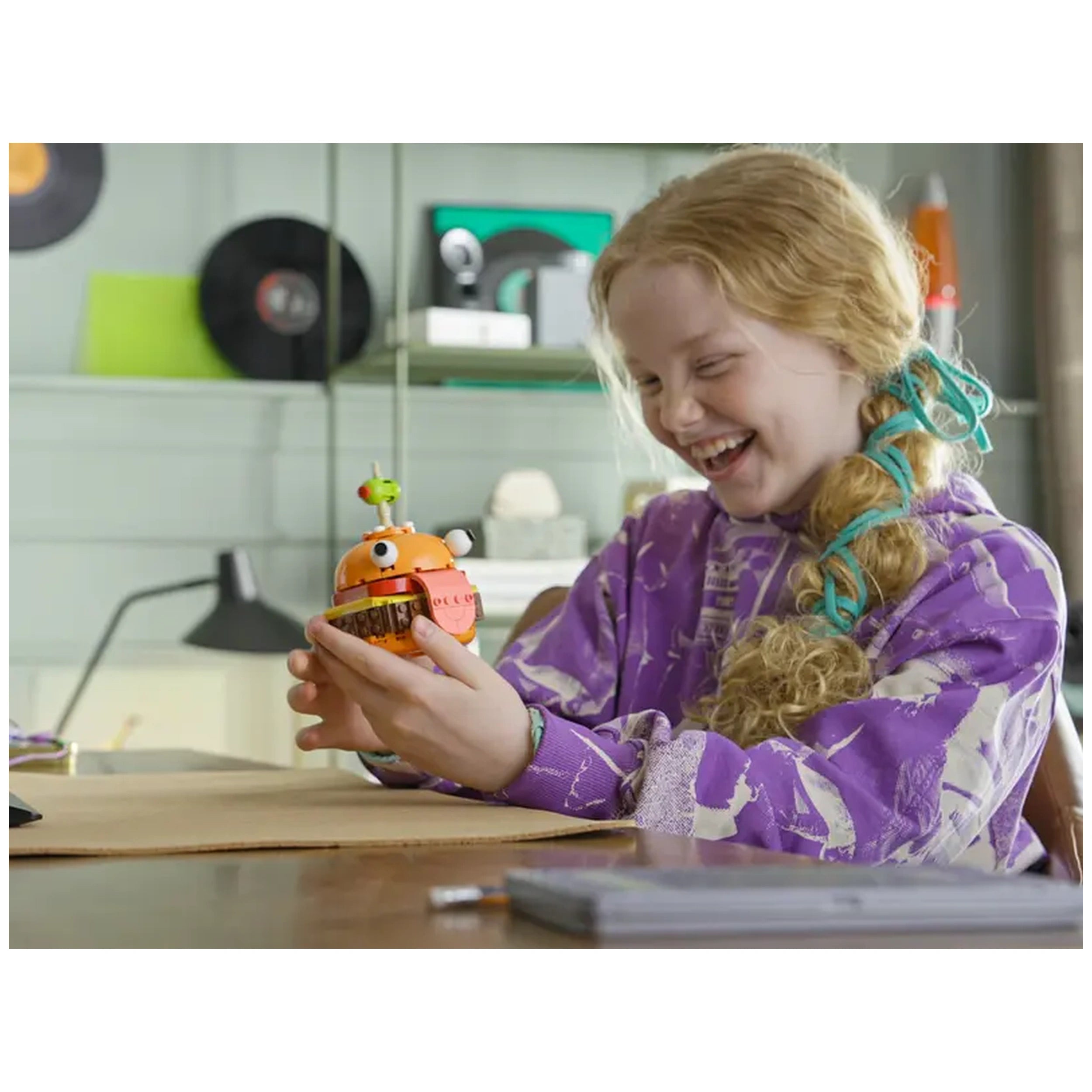 A happy child with long, wavy hair wearing a purple sweater holds the colorful LEGO® Fortnite Durrr Burger toy, smiling as she examines it. The background features green and black decor, creating a playful atmosphere.