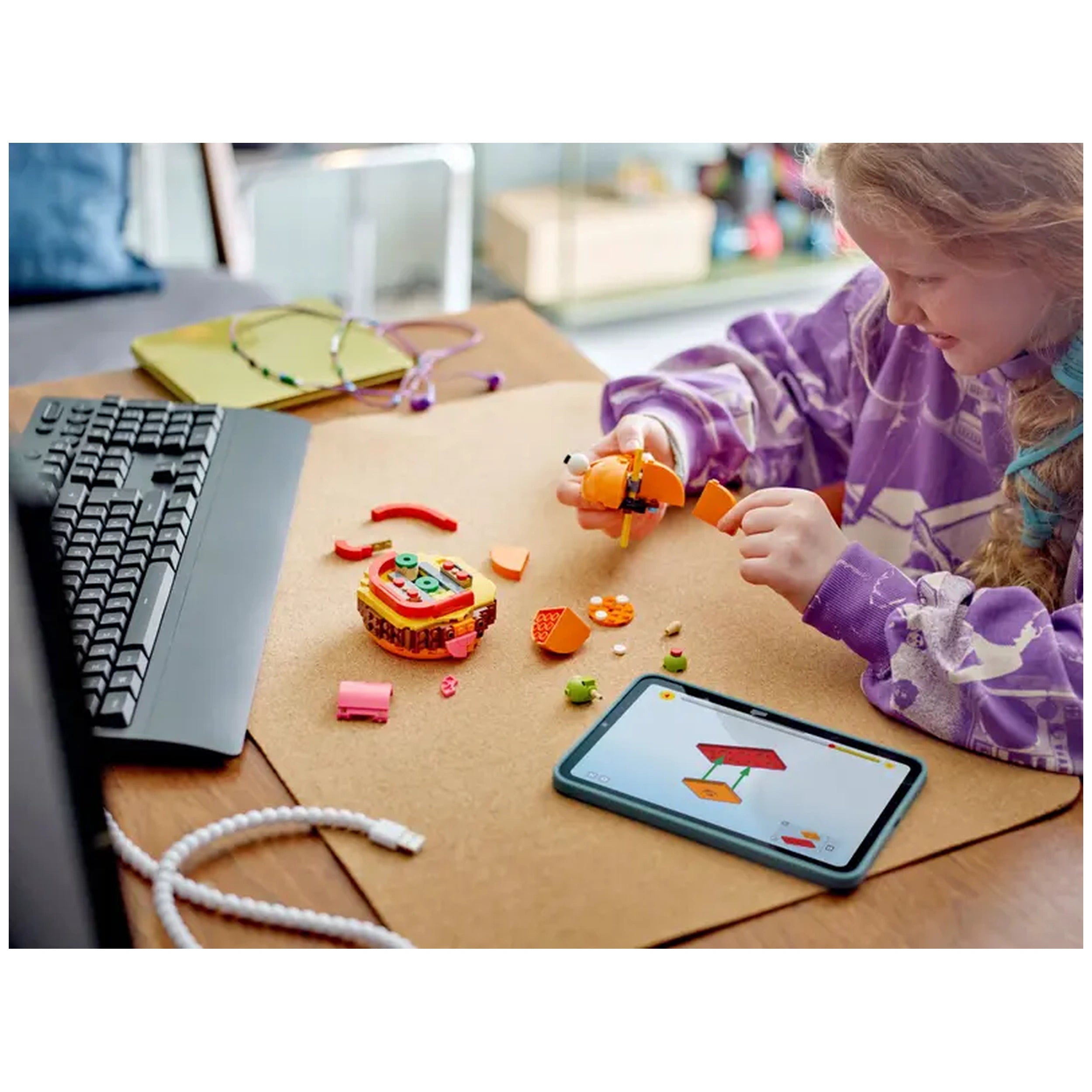 A child with curly hair wears a purple sweatshirt while assembling the LEGO® Fortnite Durrr Burger set on a cork surface. The vibrant burger pieces, including orange and green elements, are scattered around, and a tablet displays building instructions. A keyboard is visible to the left.