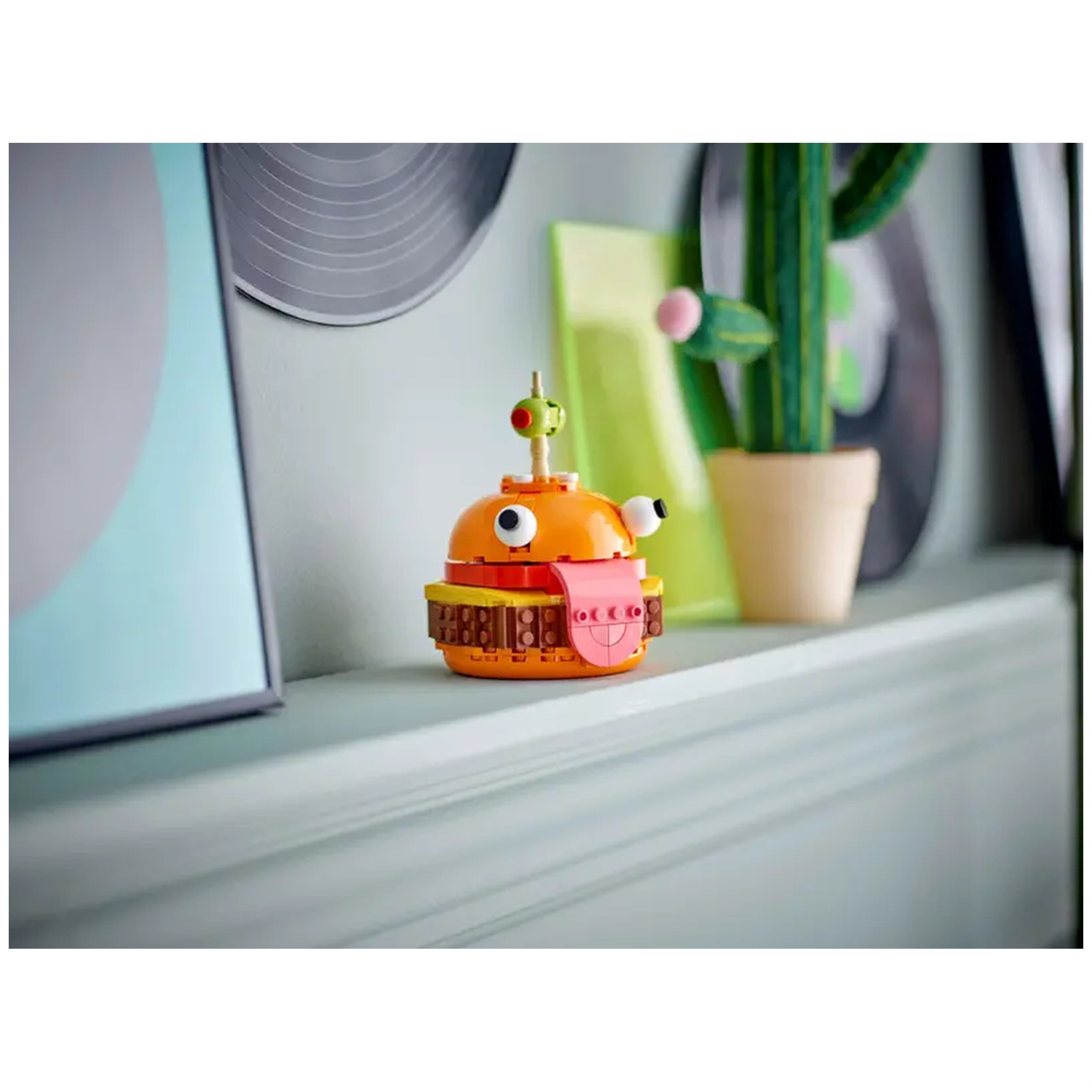 LEGO® Fortnite Durrr Burger model in vibrant orange, featuring expressive eyes and a playful tongue, displayed on a shelf among framed art and a small cactus.
