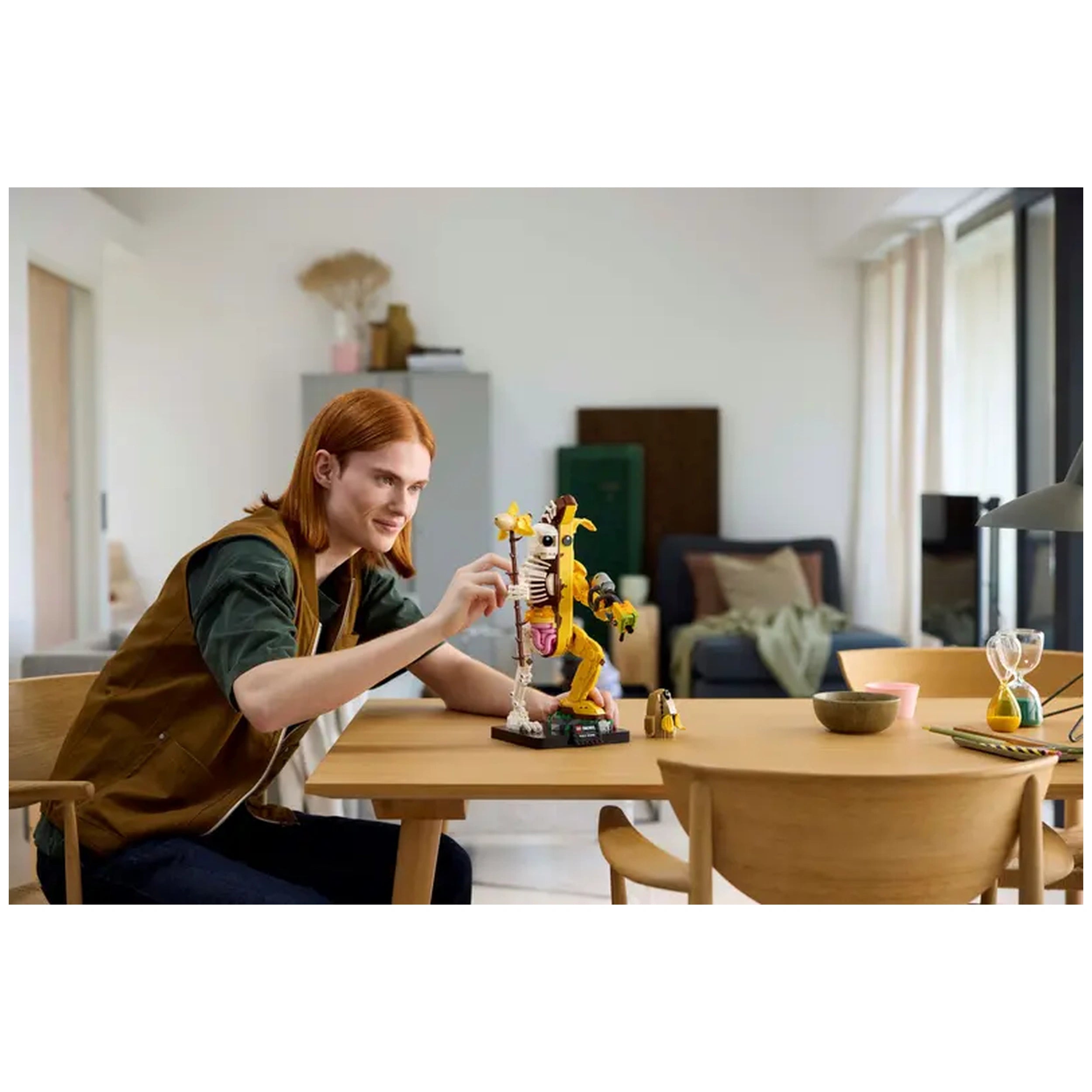 A young adult with red hair joyfully assembles the LEGO Fortnite Peely Bone construction set on a wooden table in a modern living space. The colorful figure stands tall, showcasing its playful banana and skeleton design.