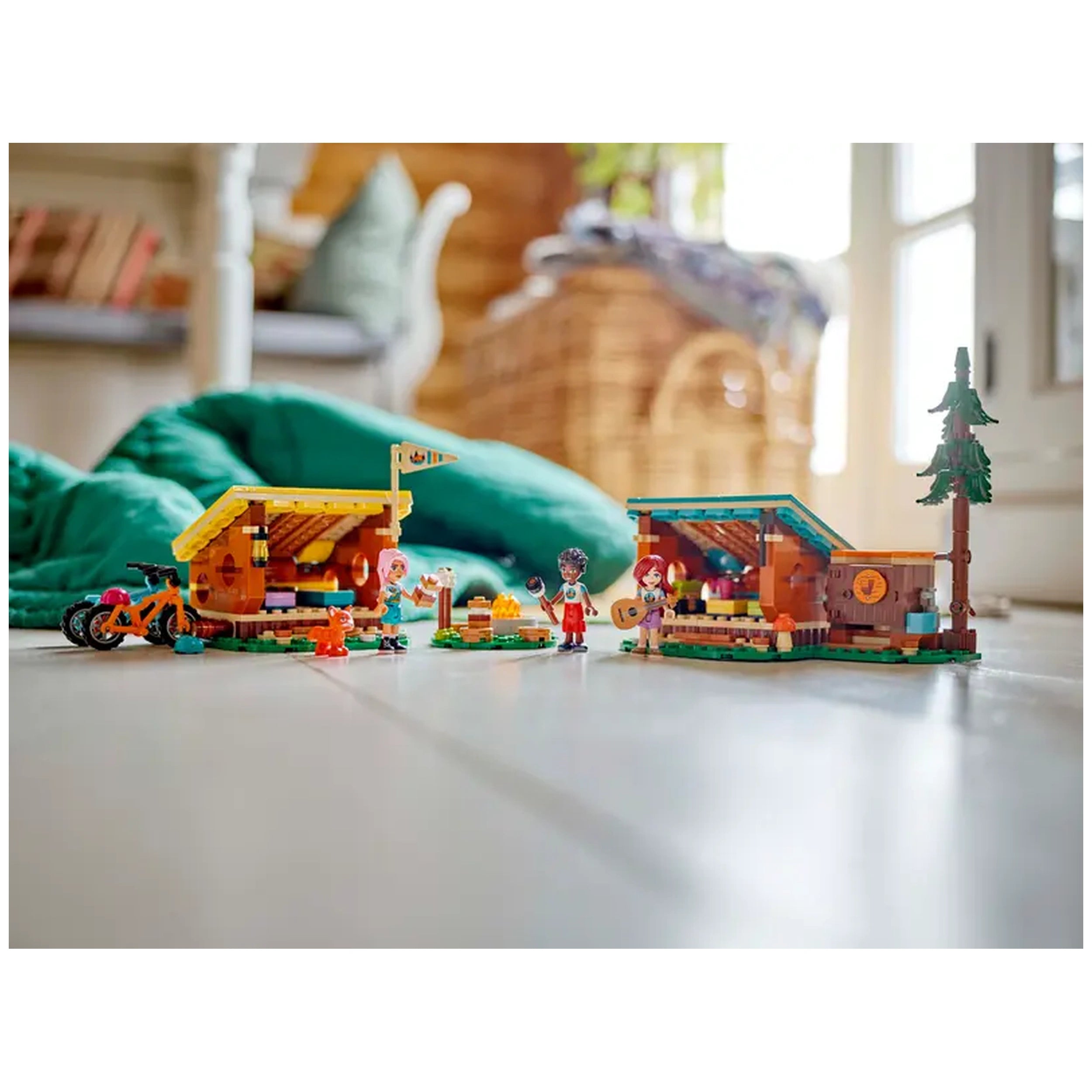 Three LEGO® Friends mini-dolls engage in a camping scene with two cozy cabins, a campfire, and mountain bikes on a wooden floor. A charming fox character adds to the adventure.