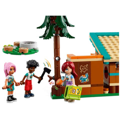 Three mini-dolls at a LEGO® Friends Adventure Camp scene, one wielding an axe near a tree, while another holds a green backpack, smiling. Two buildable cabins with colorful roofs and outdoor accessories complete the whimsical camping atmosphere.