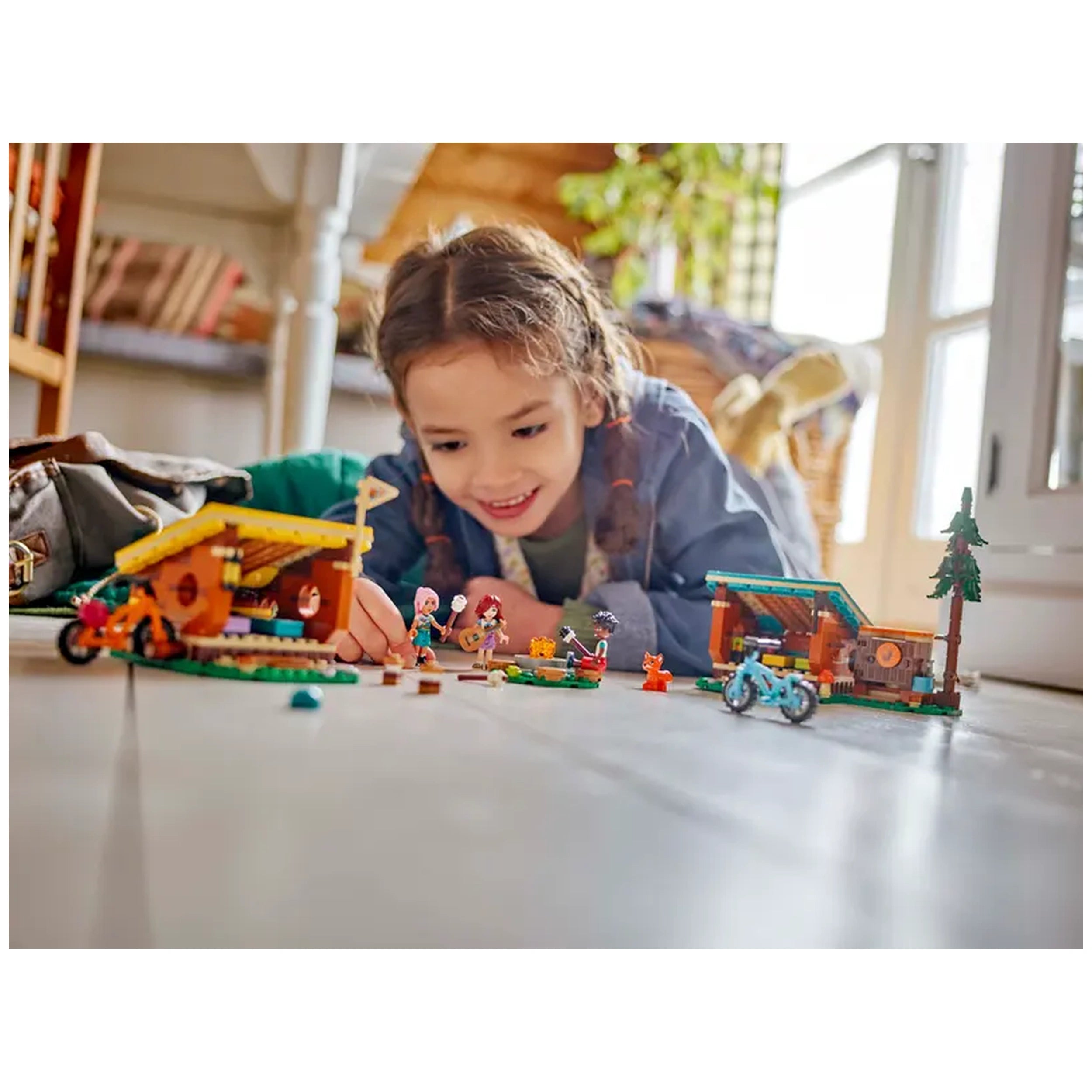 A child joyfully interacts with LEGO Friends Adventure Camp Cozy Cabins set, featuring two cabins, mini-dolls, and camping accessories on a wooden floor.