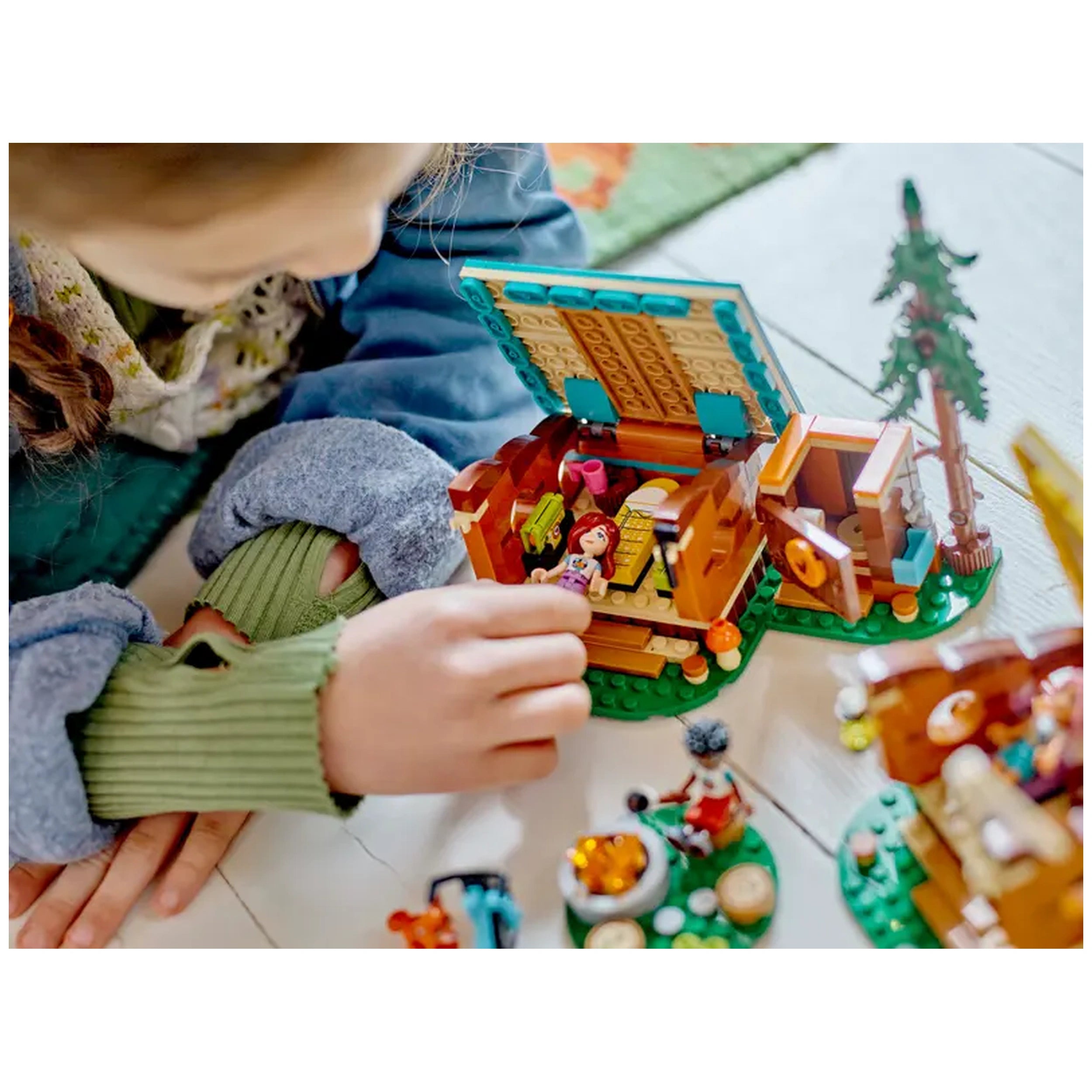 A child interacts with the LEGO® Friends Adventure Camp Cozy Cabins set, exploring a cabin with mini-dolls and camping accessories.