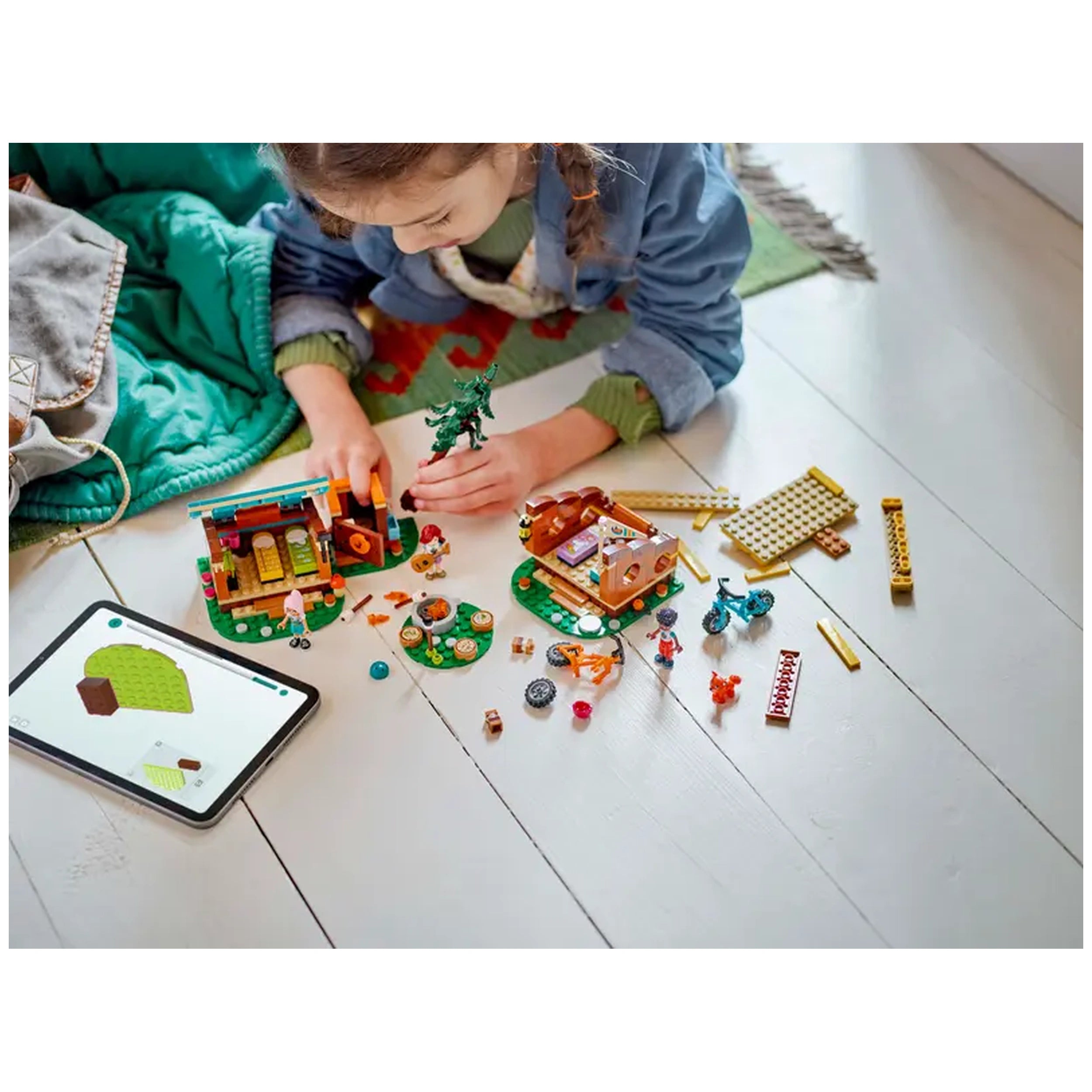 A child builds with the LEGO® Friends Adventure Camp Cozy Cabins set, featuring two colorful cabins, mini-dolls, and camping accessories on a wooden floor. An instructional tablet displays a construction guide nearby.