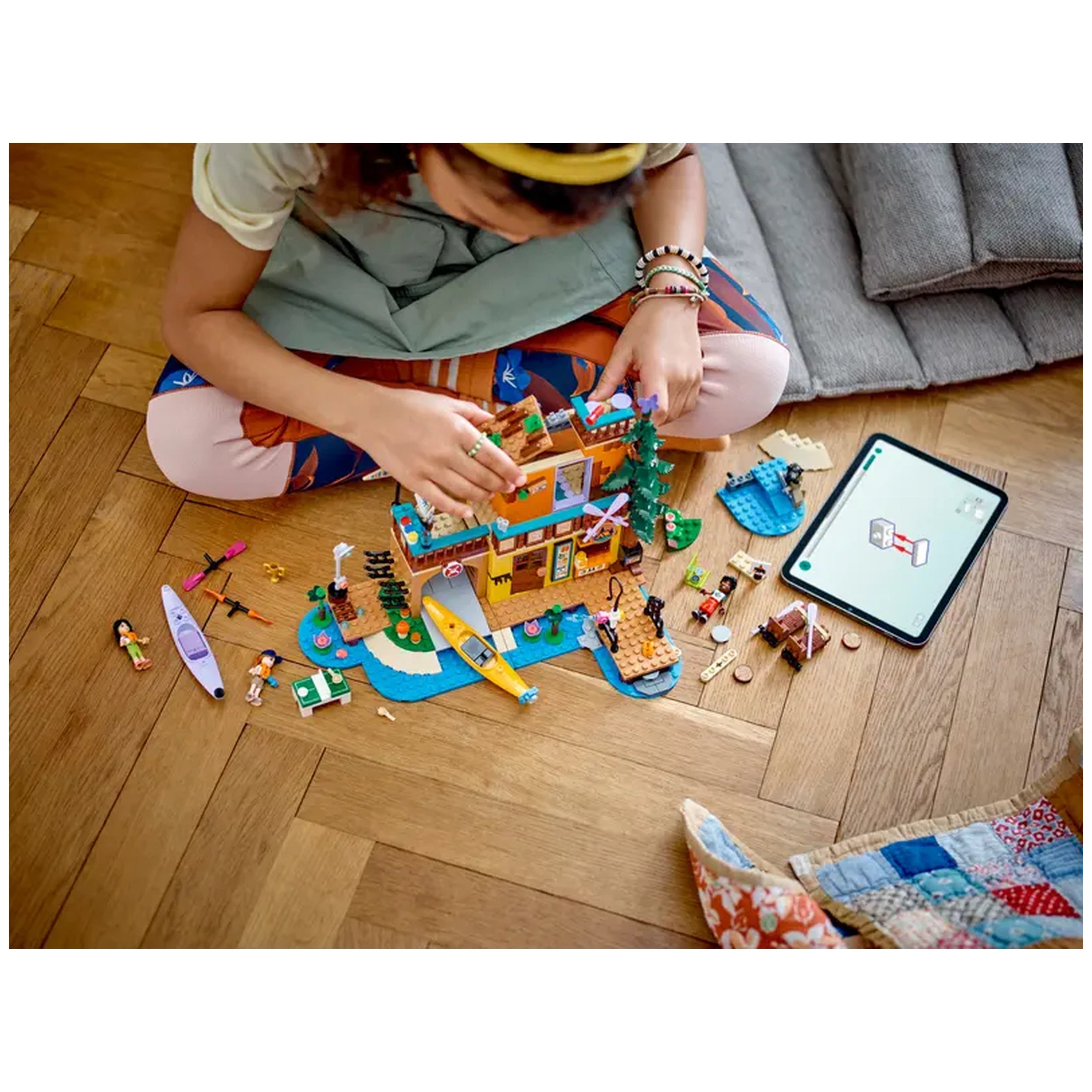 A child assembles the LEGO Friends Adventure Camp Water Sports set on a wooden floor, surrounded by colorful mini-dolls, kayaks, and building pieces, with a tablet displaying instructions nearby.