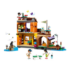 Colorful LEGO Friends Adventure Camp Water Sports set shows two mini-dolls engaged in activities outside a vibrant building. A monkey sits on a rock nearby, while butterflies float above. The scene features kayaks, a dock, and playful outdoor elements, inviting imaginative adventures.