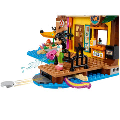 A LEGO Friends mini-doll stands on a wooden dock, holding a pink water toy, ready for fun. Behind her, colorful water sports gear is displayed, and kayaks are visible. The cheerful setup includes a vibrant cabin decorated with a life preserver and outdoor-themed items, inviting playful adventures.