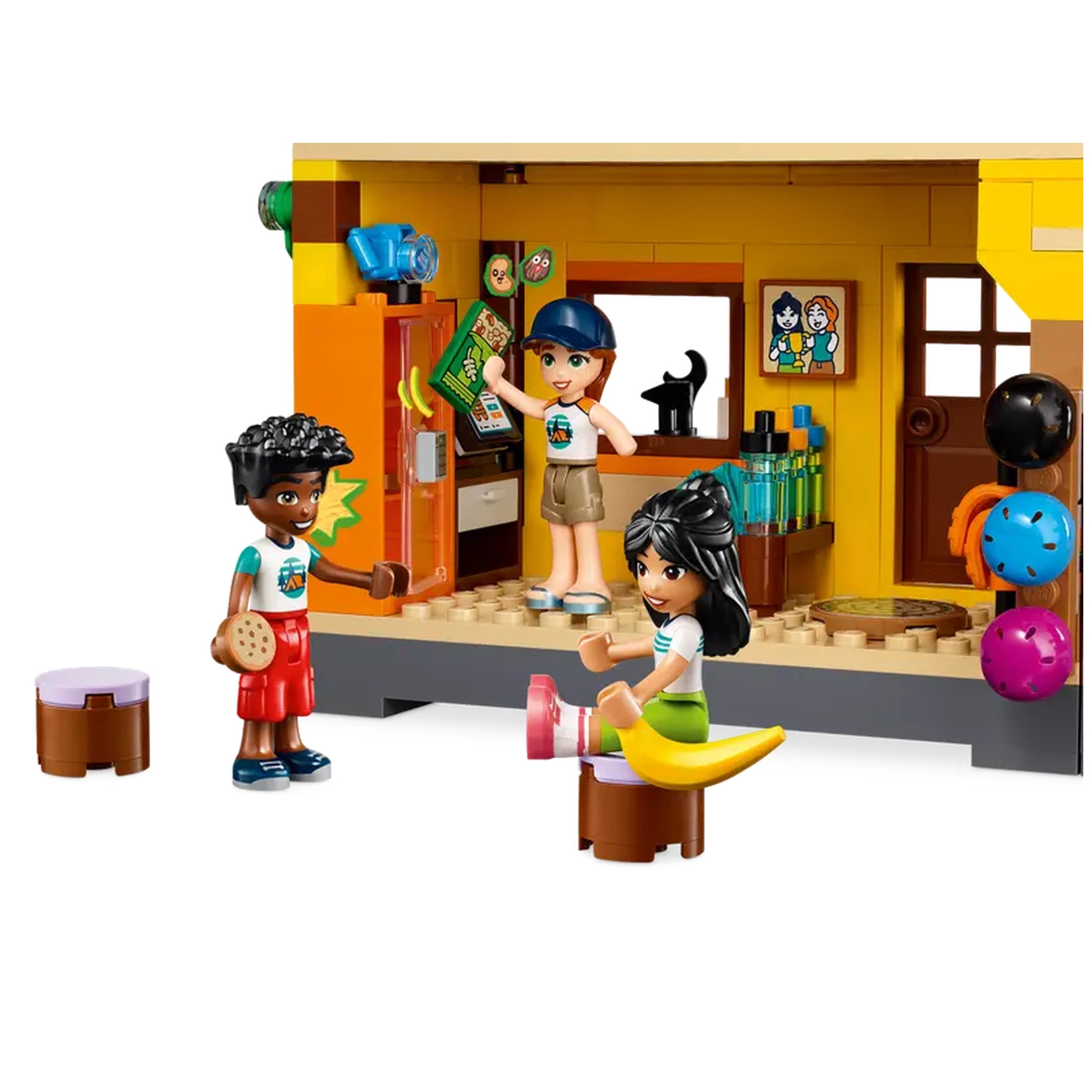 Three LEGO Friends mini-dolls engage playfully in a colorful indoor setting, with one doll retrieving a game box from a refrigerator, another holding a cookie, and a third sitting on a yellow banana-shaped seat, surrounded by playful decor and sports equipment.