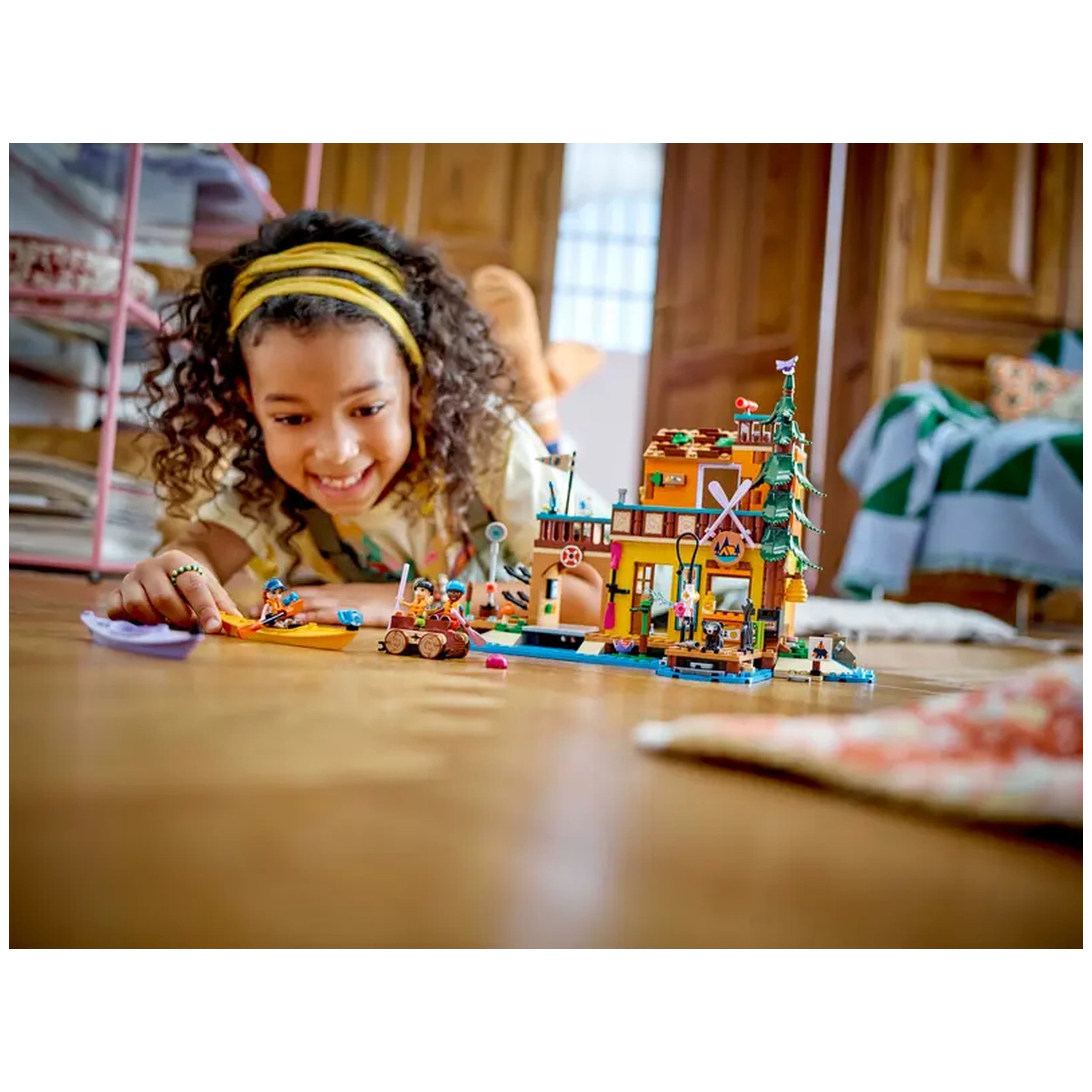 A young girl with curly hair smiles while playing on the floor, engaged with the LEGO Friends Adventure Camp Water Sports set. The colorful construction depicts a building, two kayaks, and mini-dolls, inviting creativity and outdoor adventures.