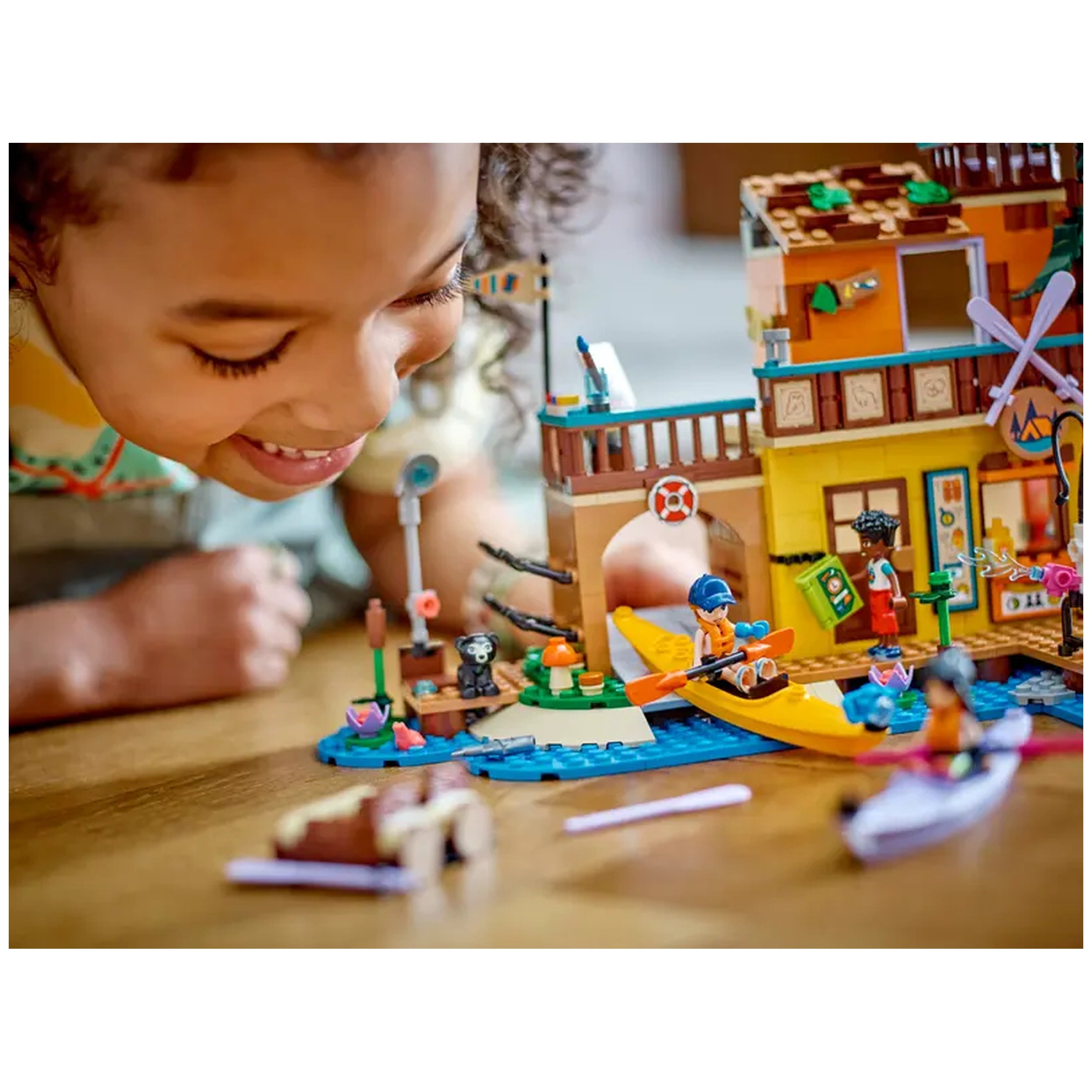 A child smiles while playing with the LEGO Friends Adventure Camp Water Sports set, featuring mini-dolls, kayaks, and a colorful dock.