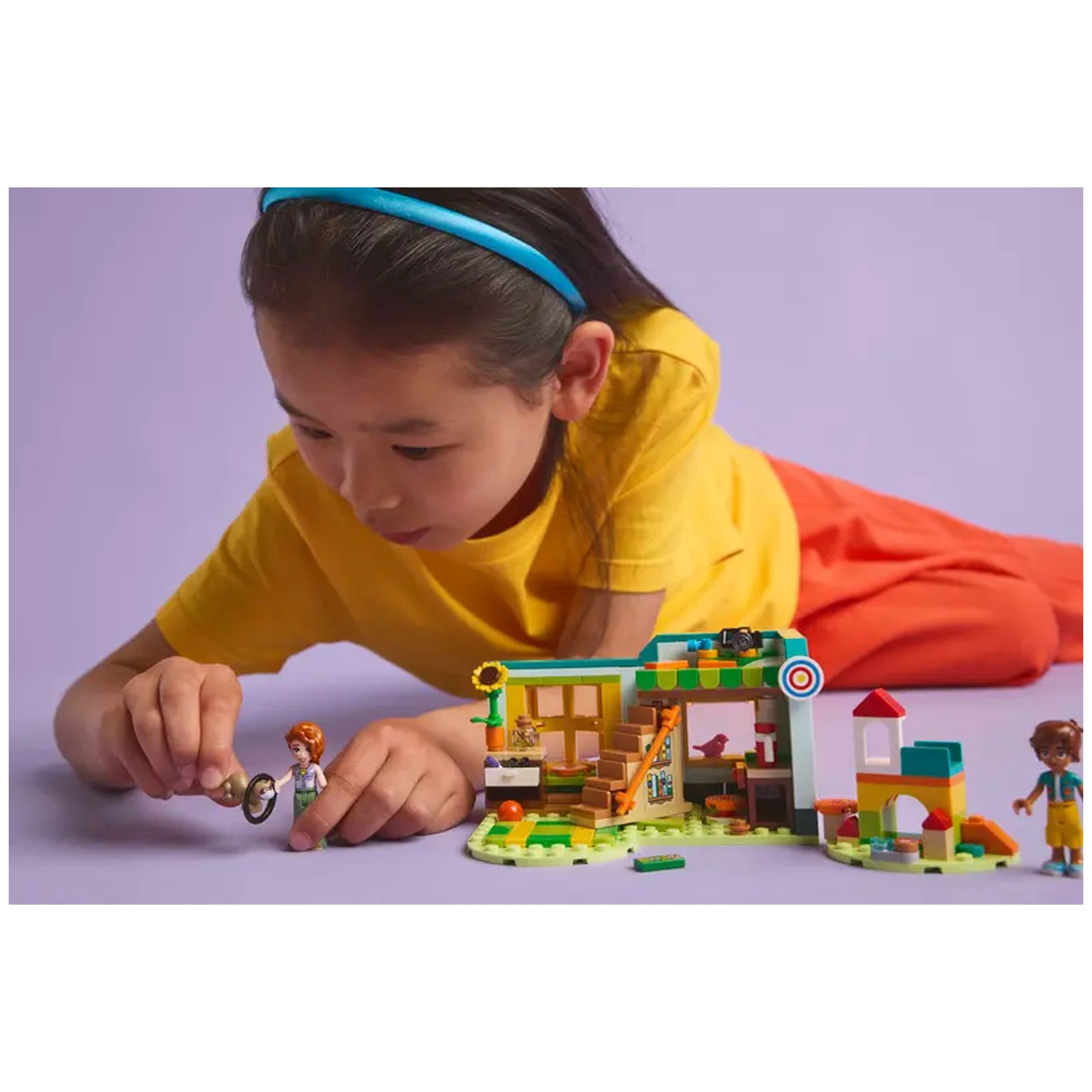 A young girl in a yellow shirt and blue headband leans over a LEGO® Friends set, engaging with a mini-doll in an autumn-themed room scene, featuring colorful accessories and a playful environment.