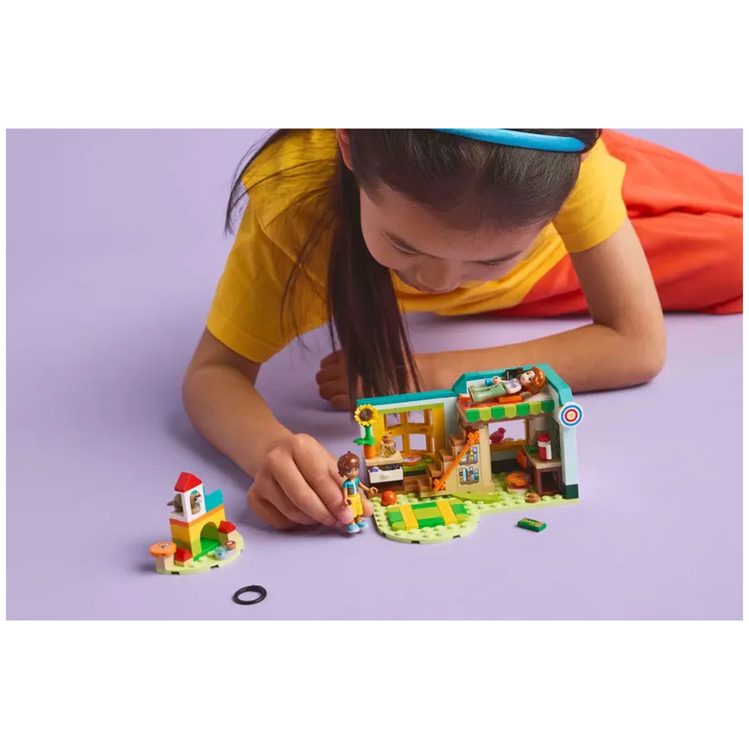 A child in a yellow shirt plays with the LEGO® Friends Autumn's Room set, which features a colorful interior scene. In the foreground, she interacts with a mini-doll, while the buildable room displays vibrant details, including a sunflower, a staircase, and various accessories. A small structure with a red roof sits nearby on a purple surface. The scene highlights creativity and imaginative play.