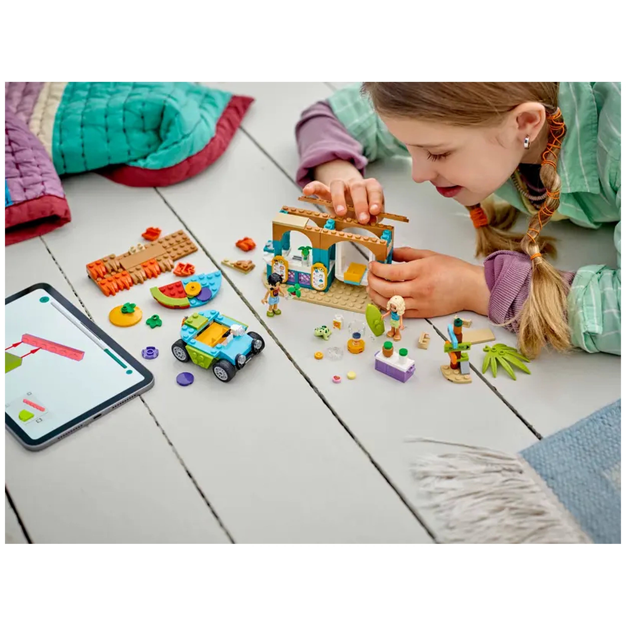 A child actively builds the Lego Friends Beach Smoothie Stand, featuring colorful bricks, mini-dolls, and a beach buggy on a light wooden floor.