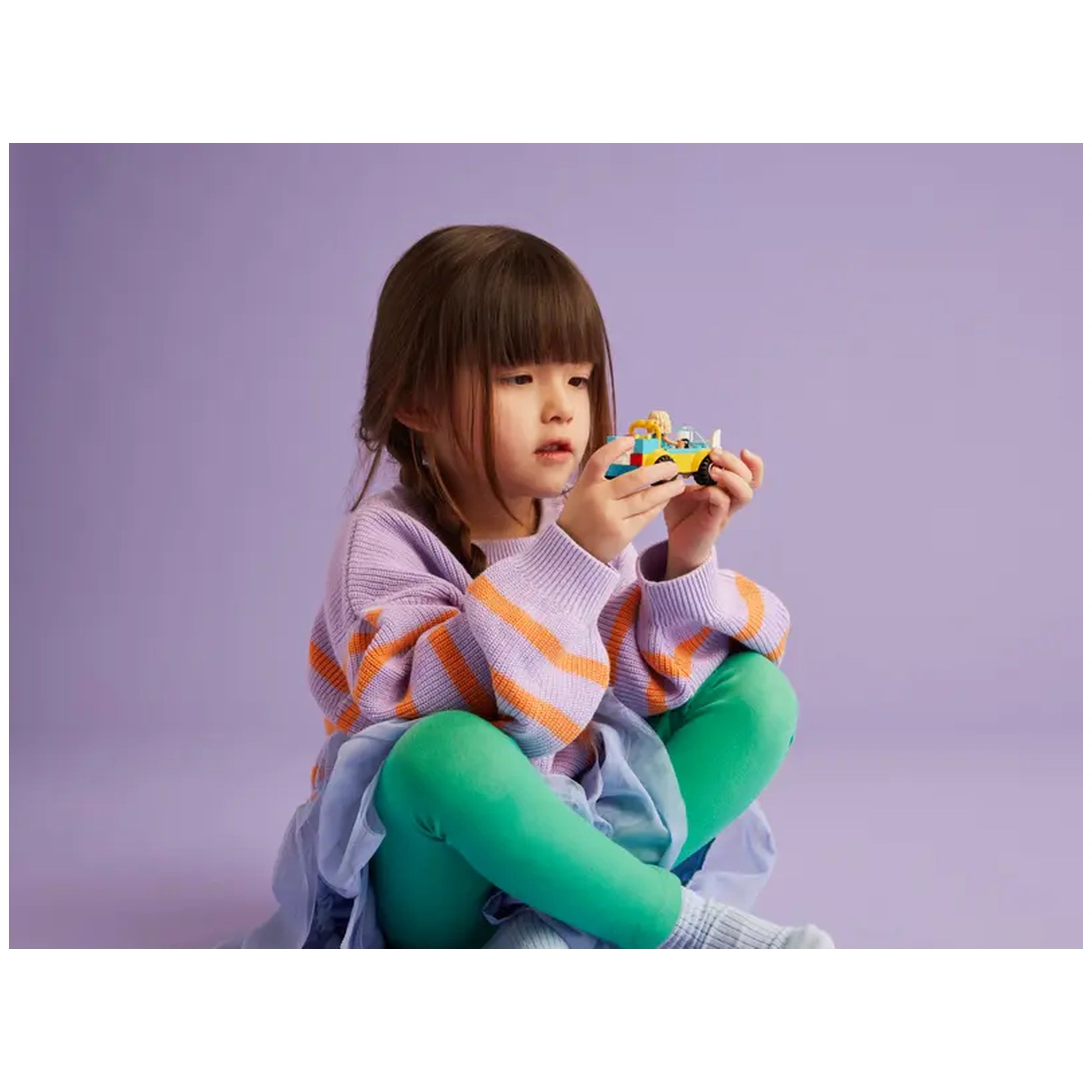 A young girl with brown hair sits cross-legged on a pastel purple background, holding a colorful LEGO® Friends Dog-Grooming Car toy. She wears a light purple oversized sweater with orange stripes and bright green leggings.