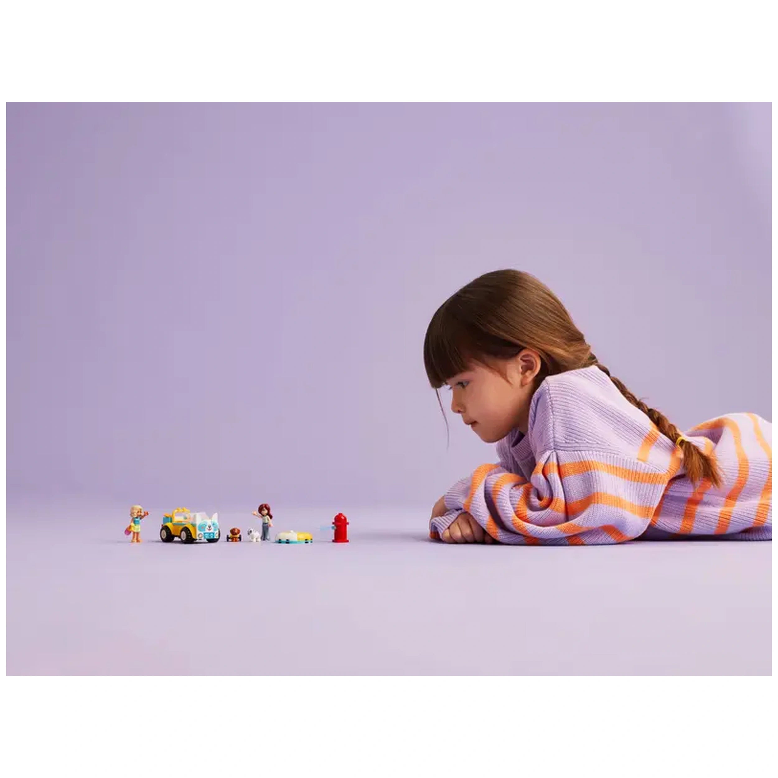 A young girl lies on her stomach, engaged with the LEGO® Friends Dog-Grooming Car set. She curiously observes two mini-dolls, a dog, and various colorful pet care accessories against a soft purple background.