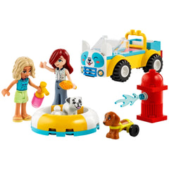 Two mini-dolls, one with curly blonde hair and one with straight red hair, are playing with a white dog in a circular pool. A colorful dog-grooming car and a red fire hydrant are nearby, with a small brown dog on a yellow cart. The scene is vibrant and playful, reflecting a fun pet care activity.
