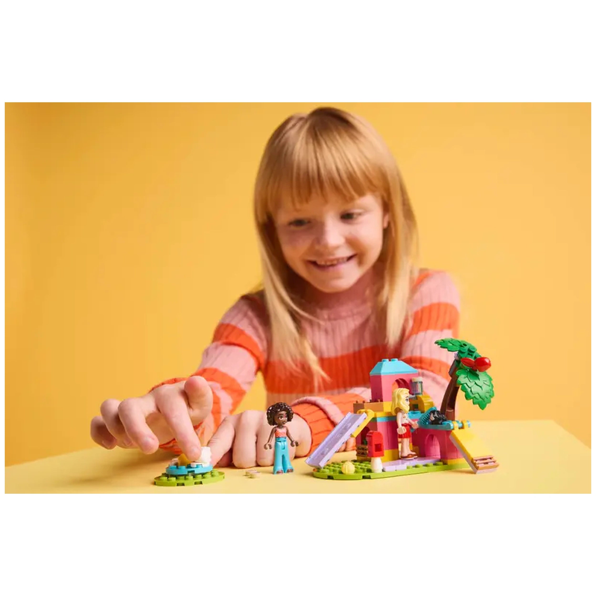 A young girl with long blonde hair smiles while interacting with the LEGO Friends Guinea Pig Playground set. She is placing a white guinea pig figure on a green base, surrounded by colorful playset elements like a slide, tree, and two minidolls. The background is a vibrant yellow, enhancing the playful atmosphere.