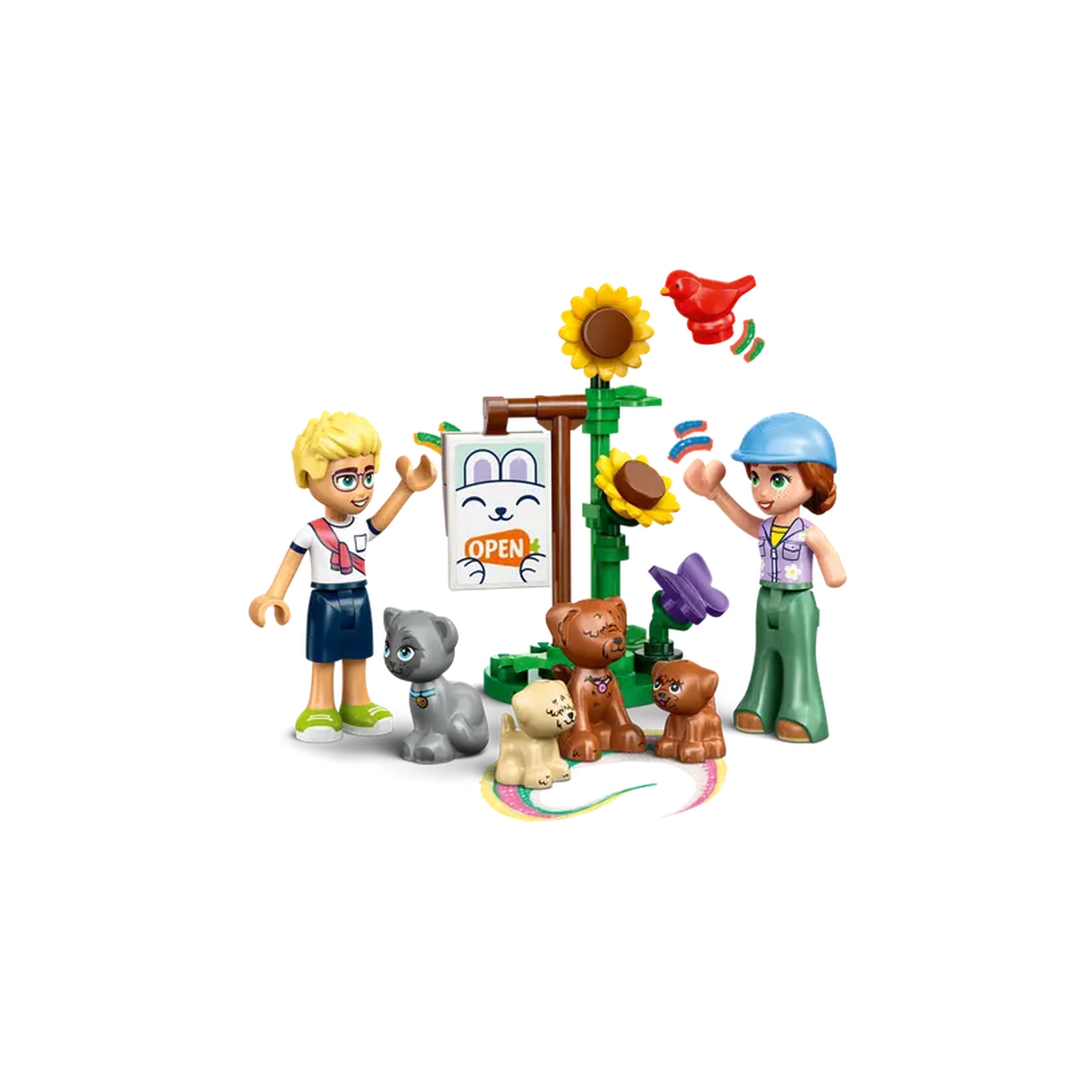 Two LEGO Friends minidolls, one with blond hair and glasses, the other wearing a blue cap, stand excitedly beside an "OPEN" sign in front of a vibrant veterinary clinic with sunflowers. Surrounding them are three playful puppies and a gray kitten, creating a lively scene of animal care and friendship.