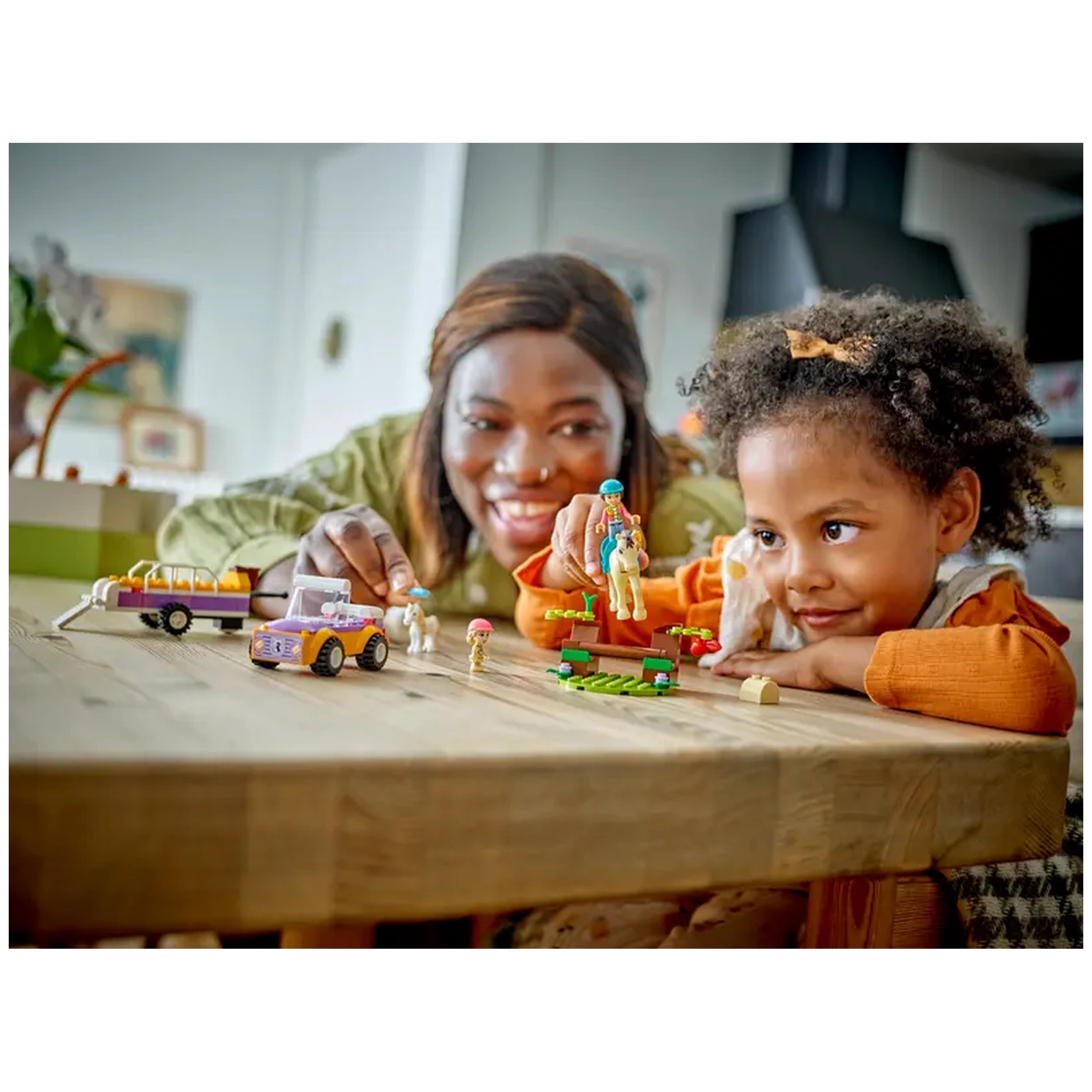 A smiling adult and child play with LEGO Friends Horse and Pony Trailer set, featuring a car, horse, and colorful accessories on a wooden table.