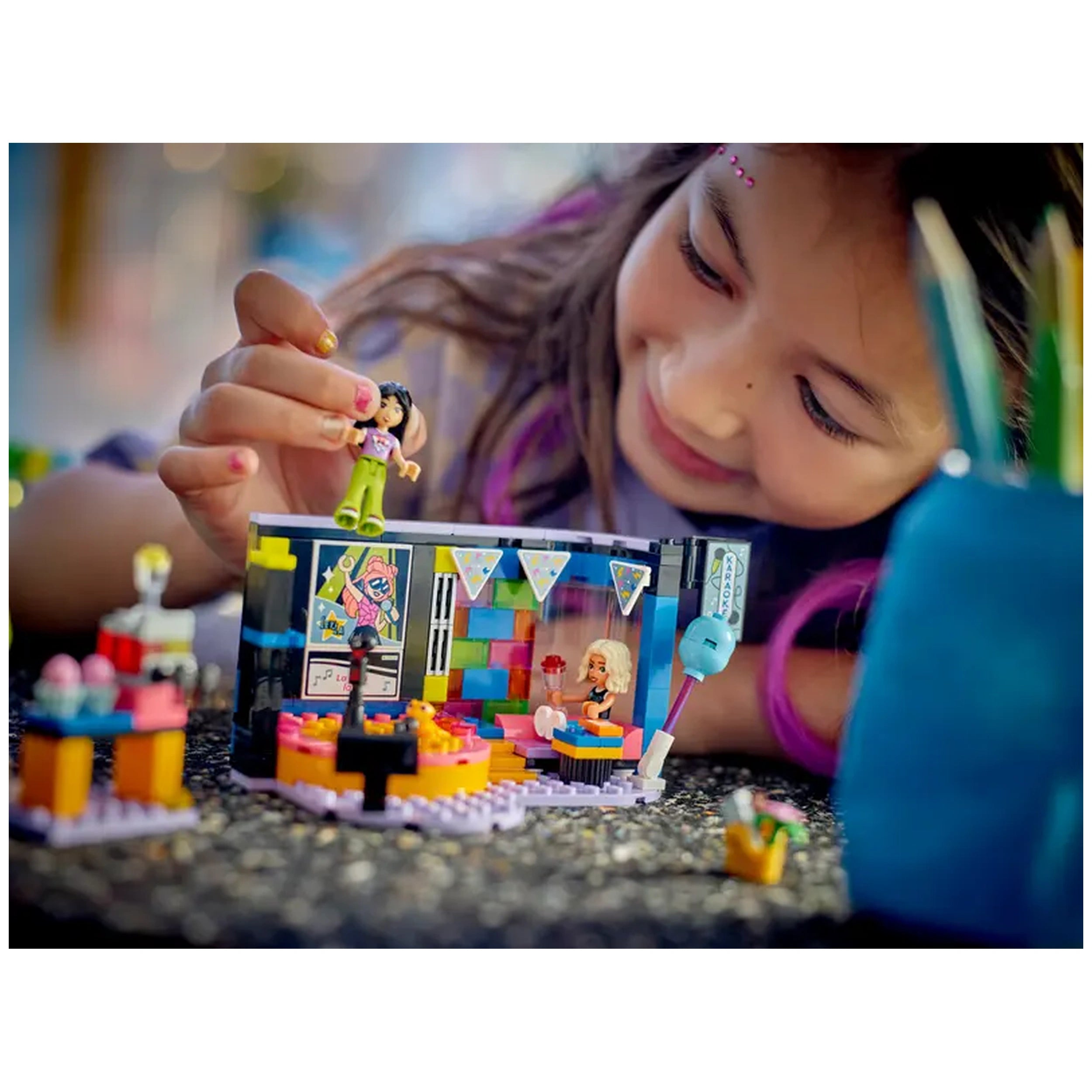 A young girl joyfully interacts with the LEGO® Friends Karaoke Music Party set, featuring colorful building elements, a revolving stage, and two mini-dolls, all surrounded by vibrant decorations and a cozy atmosphere.