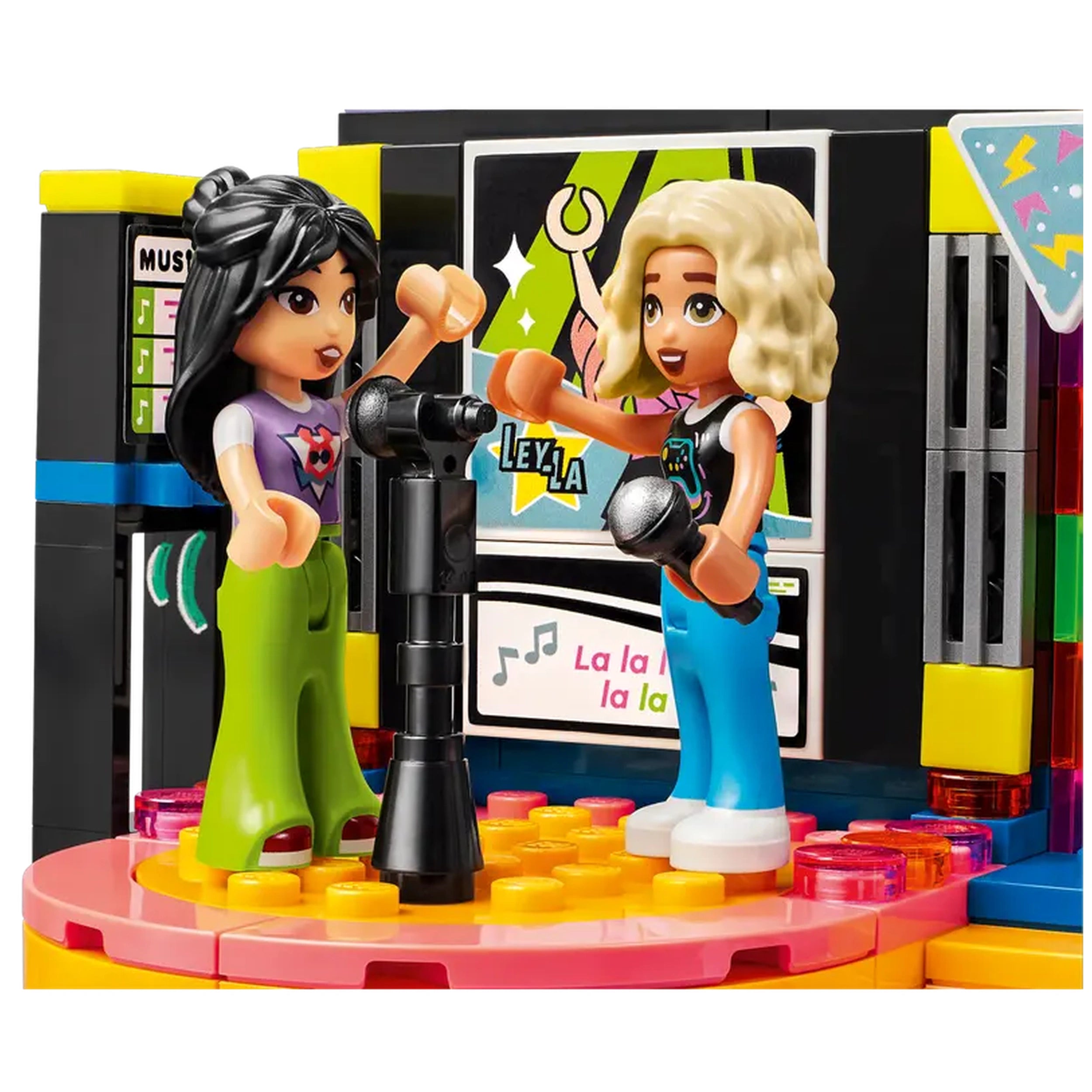 Two LEGO® Friends characters singing at a karaoke stage, one with black hair in a pink shirt and green pants, the other with blonde hair in a black top and blue pants, holding microphones. The colorful stage features a display screen and vibrant lights, creating a lively party atmosphere.