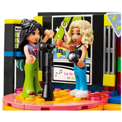 Two LEGO® Friends characters singing at a karaoke stage, one with black hair in a pink shirt and green pants, the other with blonde hair in a black top and blue pants, holding microphones. The colorful stage features a display screen and vibrant lights, creating a lively party atmosphere.