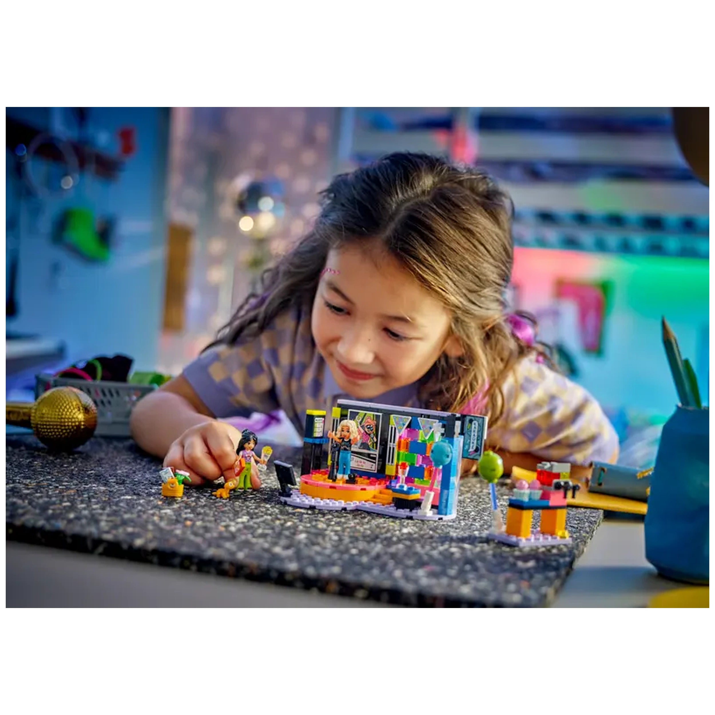 A young girl with long hair smiles as she plays with the LEGO Friends Karaoke Music Party set, featuring a revolving stage, microphones, and colorful decorations. The scene is set on a textured black surface, with a bright background suggesting an exciting party atmosphere.