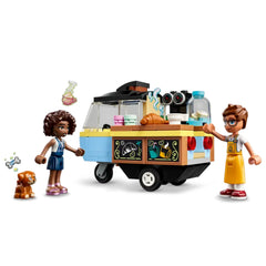Two LEGO® mini-dolls, one wearing a striped dress and the other in an apron, interact by a colorful mobile bakery cart, featuring treats like croissants. A small dog sits nearby, with playful icons of pastries and steam rising from the cart.