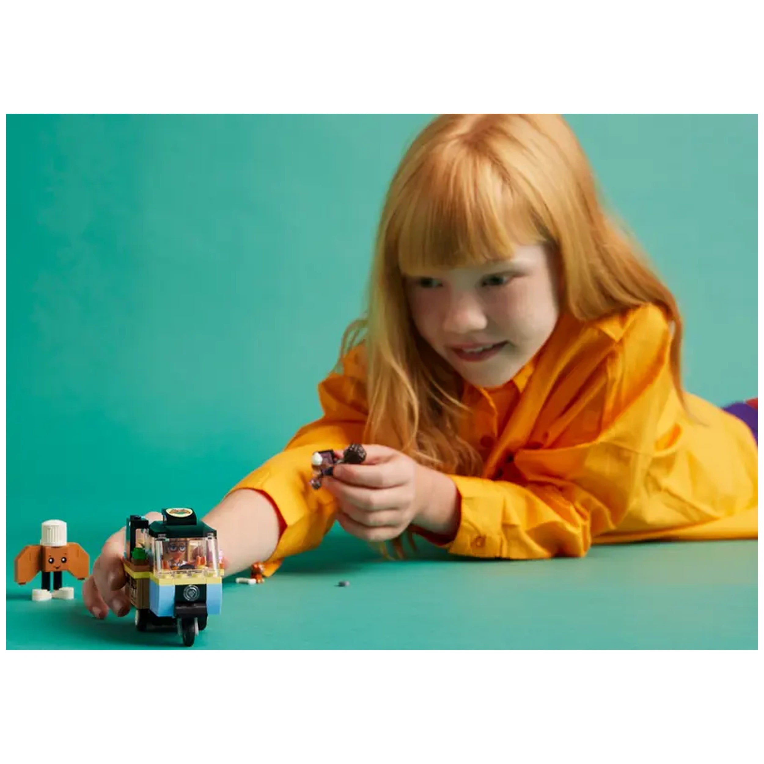 A young girl with long, red hair smiles while playing with the LEGO® Friends Mobile Bakery Food Cart set, interacting with mini-dolls and a dog figure, on a teal surface.