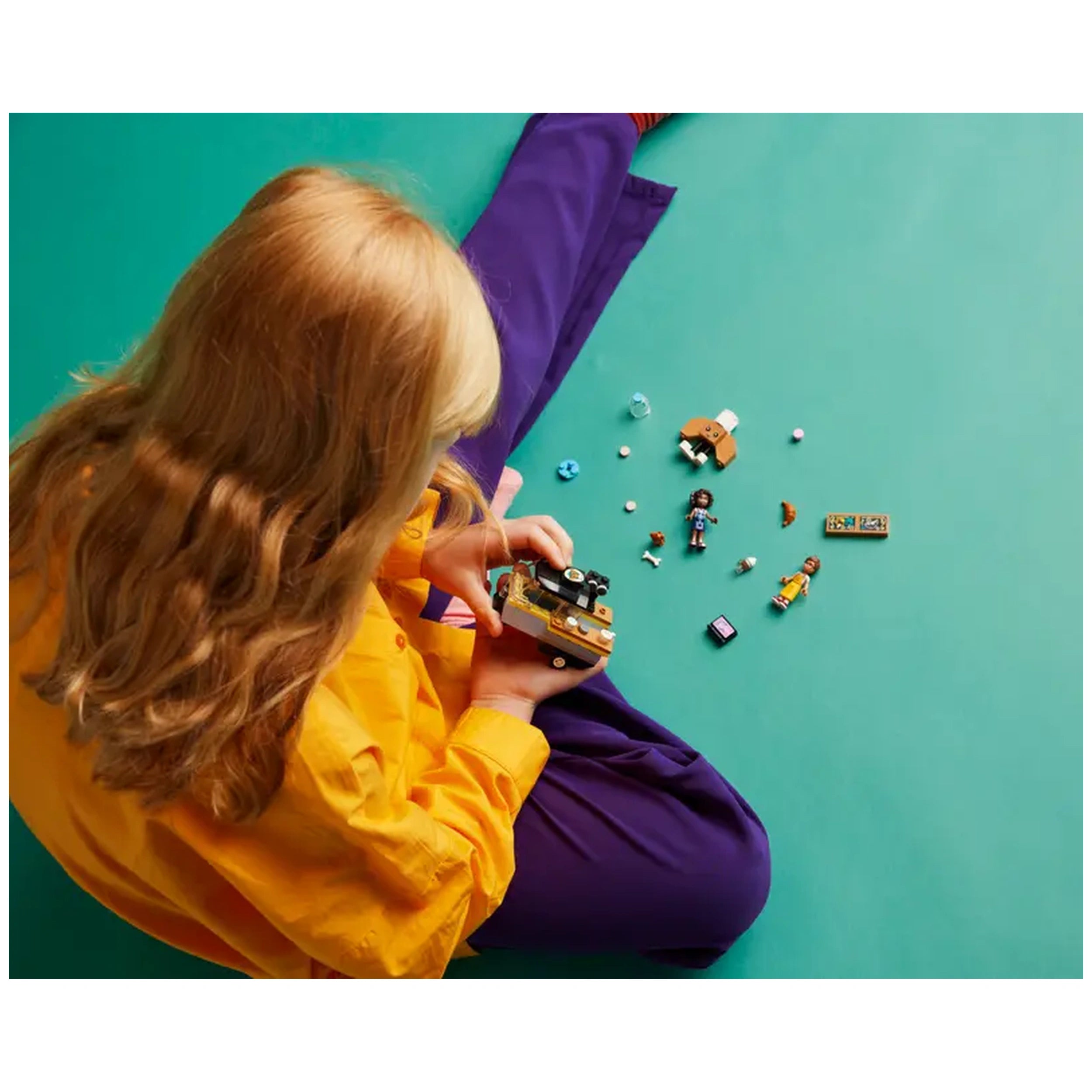 A child in a bright yellow shirt sits on a teal surface, playing with the LEGO® Friends Mobile Bakery Food Cart. Surrounding them are various colorful accessories, mini-dolls, and a dog figure, inviting imaginative play and creativity.