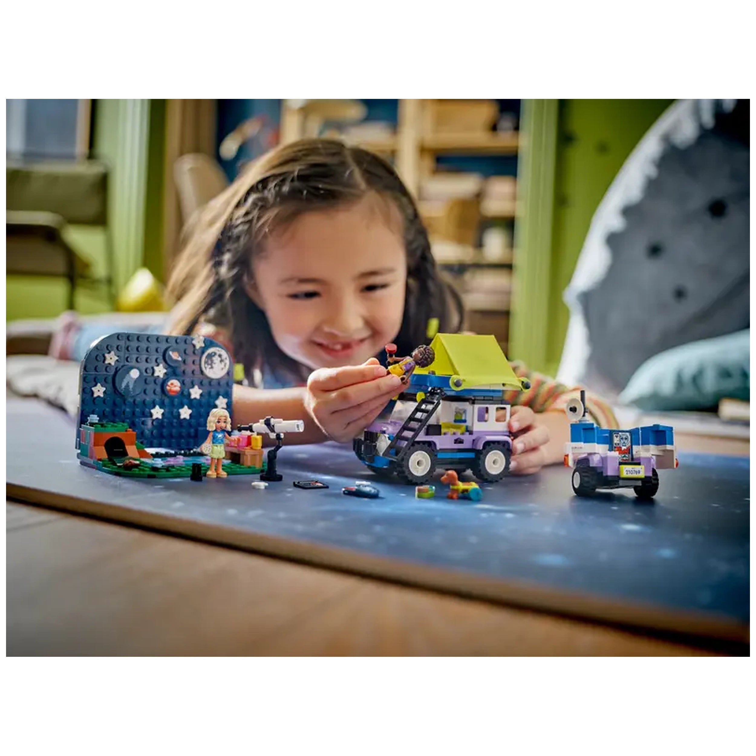 A child joyfully plays with the LEGO® Friends Stargazing Camping Vehicle set, assembling the bright 4x4 vehicle with a camping trailer and toys scattered around, including a mini-doll and a telescope, on a tabletop.
