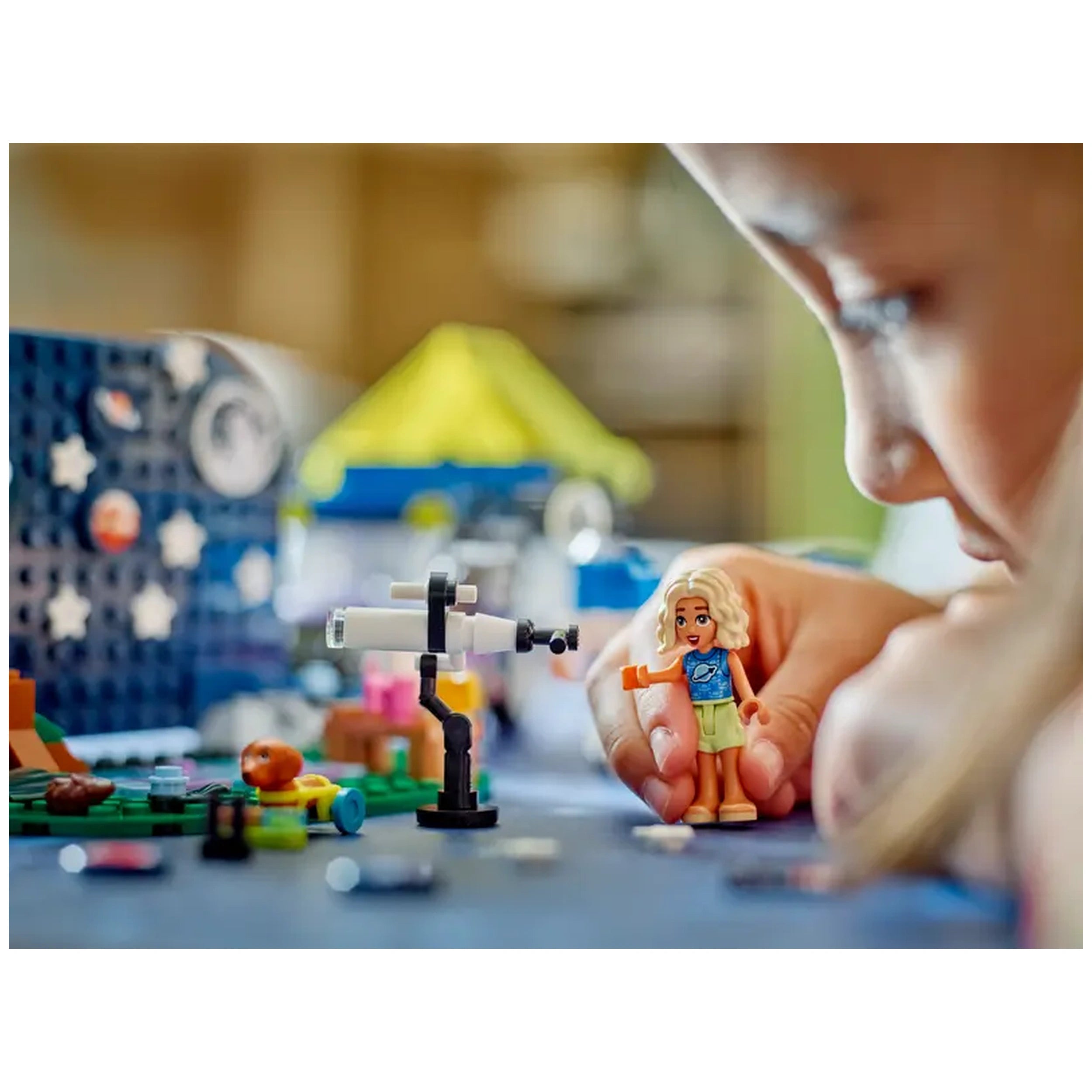 Child playing with a LEGO mini-doll in front of a toy telescope, colorful building blocks, and a camping setup with a yellow tent.