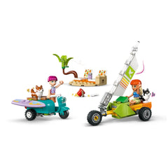 Two LEGO Friends minidolls, one with pink hair, wave from colorful scooters alongside corgi figures. A beach scene with a palm tree, lounge chairs, and snacks is nearby.