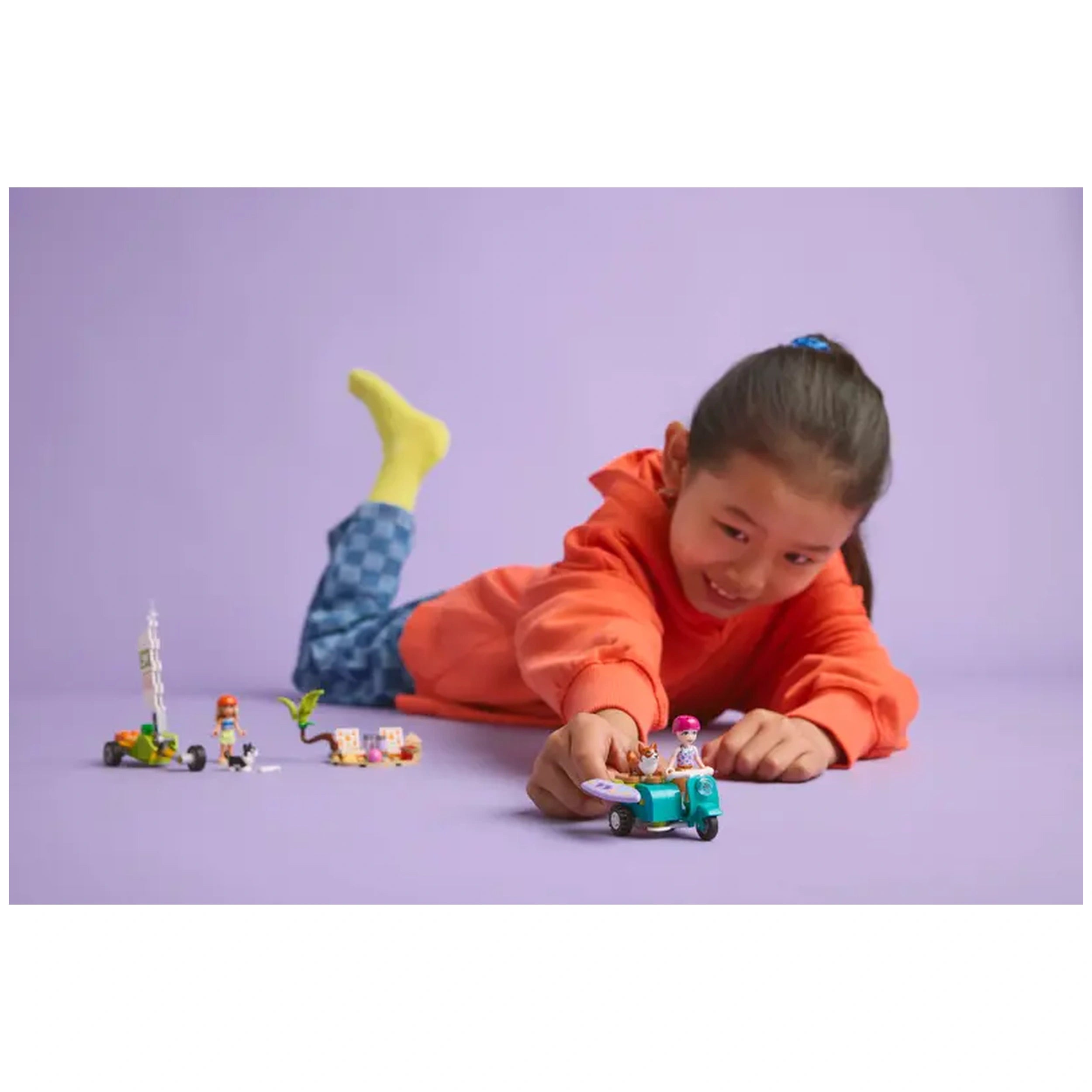 A child in an orange hoodie and blue checkered pants plays on a purple background with a LEGO Friends scooter and minidolls, smiling joyfully.