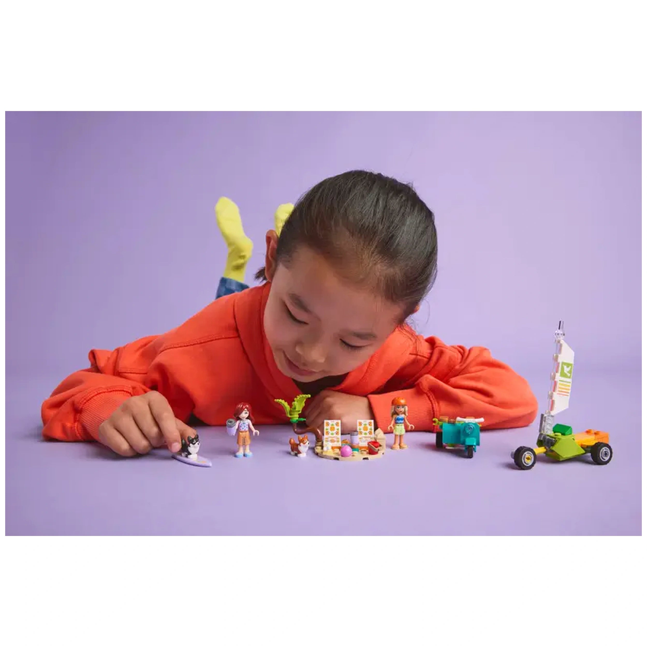 A child in a bright orange sweater leans over a colorful array of LEGO Friends figures, including two minidolls and playful accessories, on a purple background.
