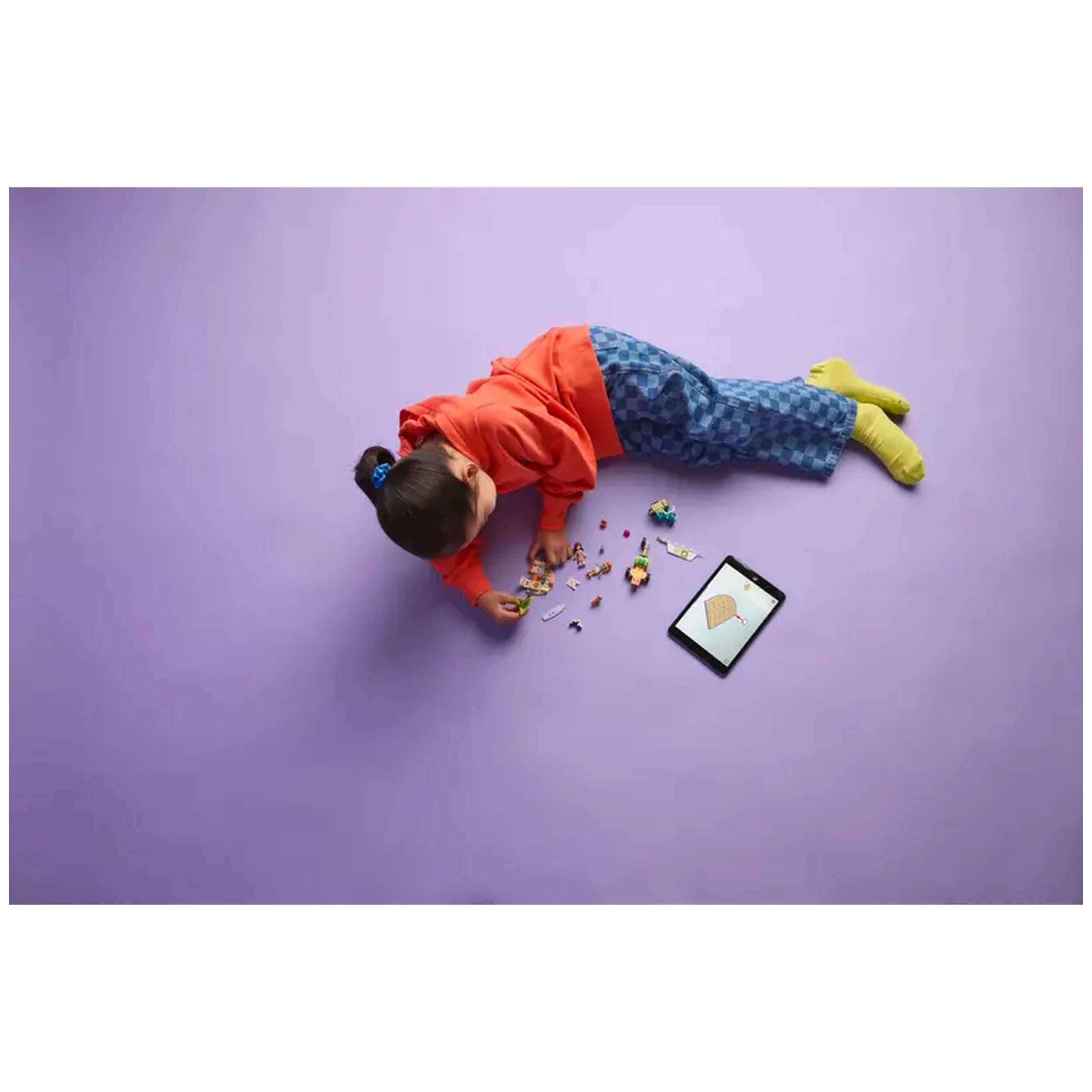 A child in a red hoodie and blue patterned pants lays on a purple floor, playing with colorful LEGO pieces and mini dolls, while an iPad displaying an ice cream cone sits nearby.
